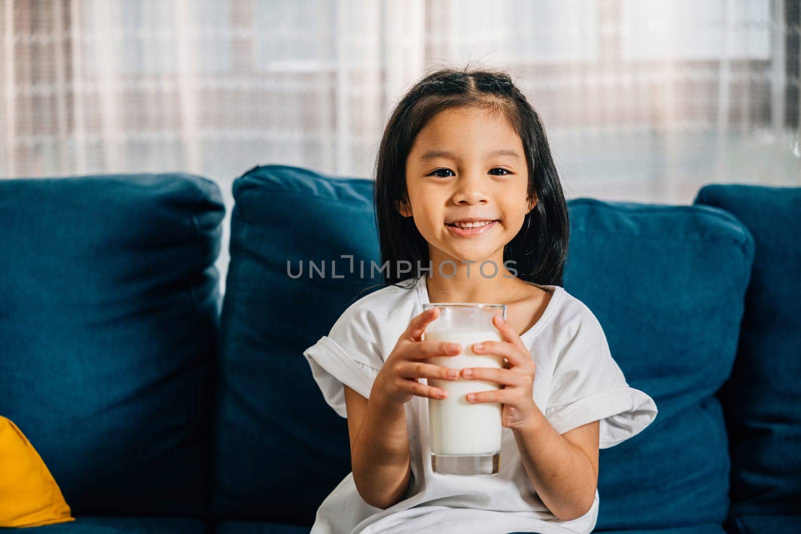 Asian child enjoys a glass of milk on the sofa their happiness and innocence shining through by Sorapop