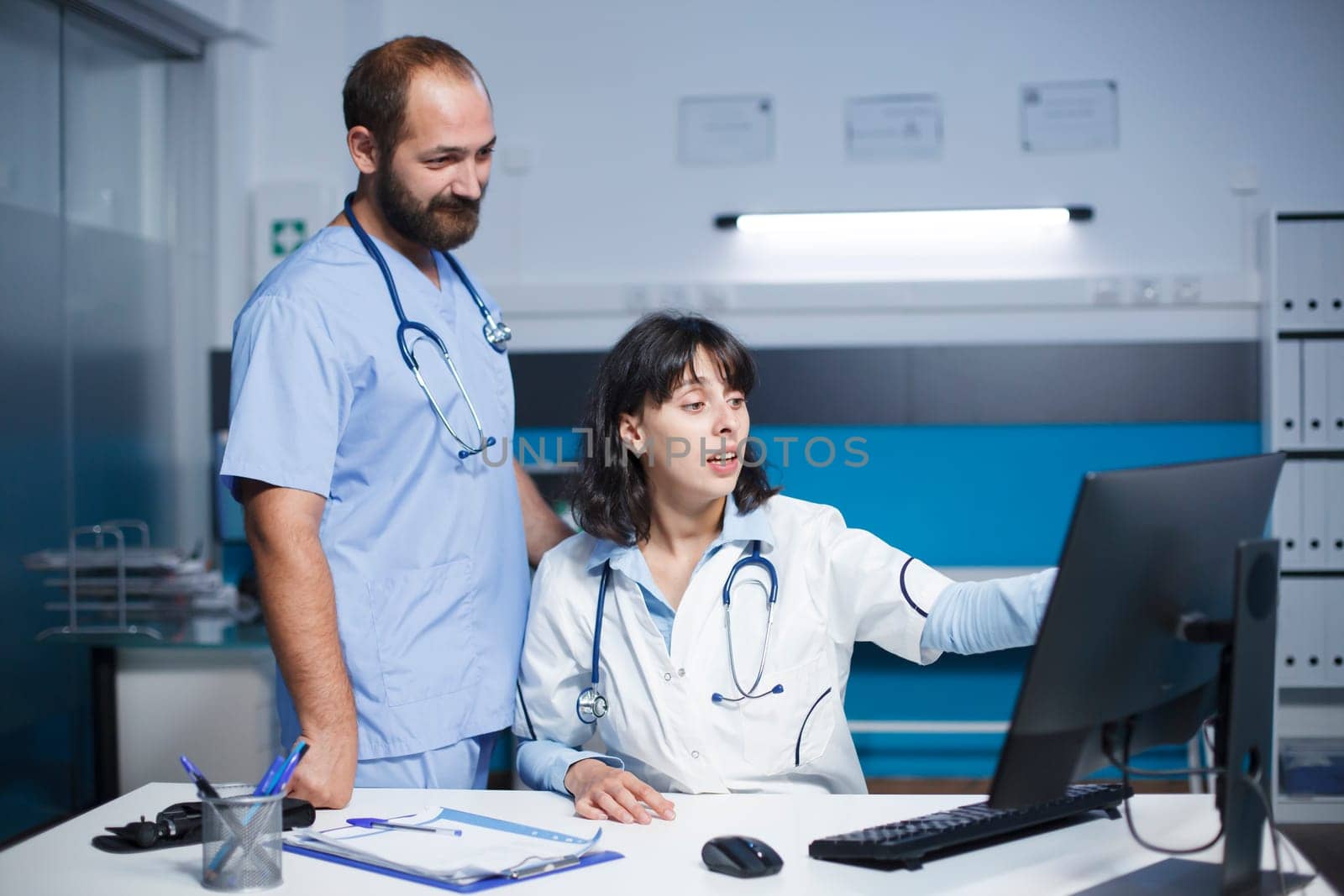 Doctor and nurse in clinic office preparing for medical consultations with patients. Their lab coats denote their profession, and the blue scrubs indicate a surgical role.