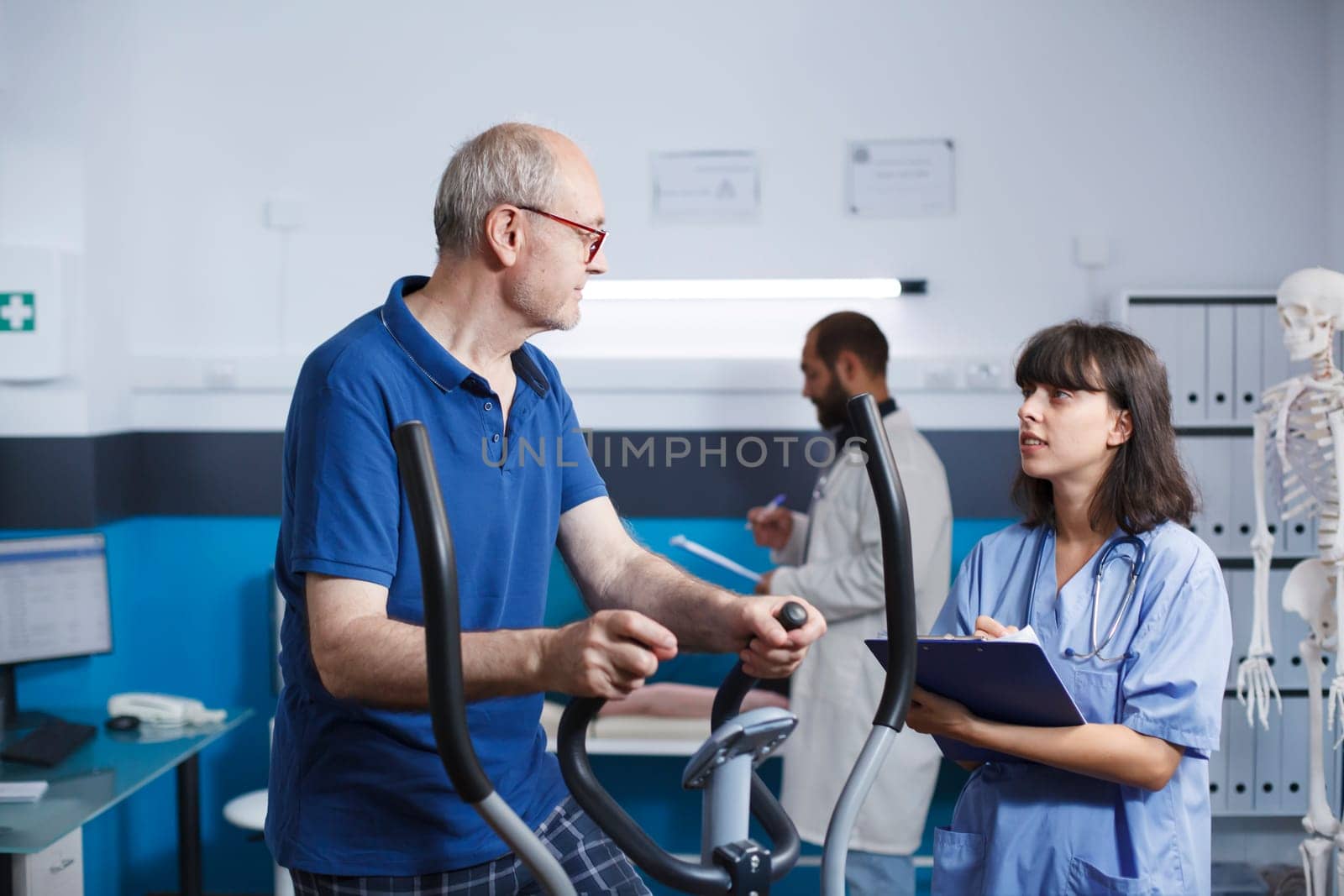 Elderly patient is seen receiving physiotherapy on stationary bike, assisted by female nurse dressed in blue scrubs. Senior man is getting rehabilitative care with help from health professional.