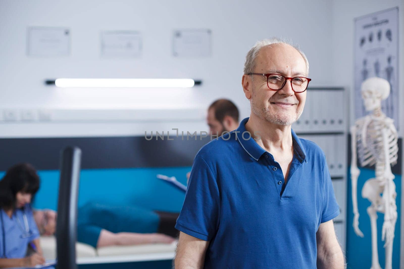 Elderly man with glasses standing in a physiotherapy facility. Retired senior patient smiling at camera as he gets ready for his recovery at clinic for rehabilitation treatment through fitness.