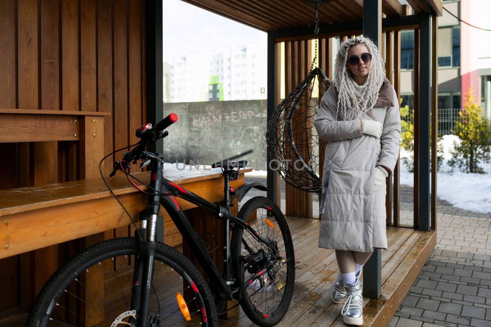 A woman is standing next to a bicycle on a porch. She is wearing casual clothing and appears to be looking out in the distance. The bike is leaning against the railing of the porch.