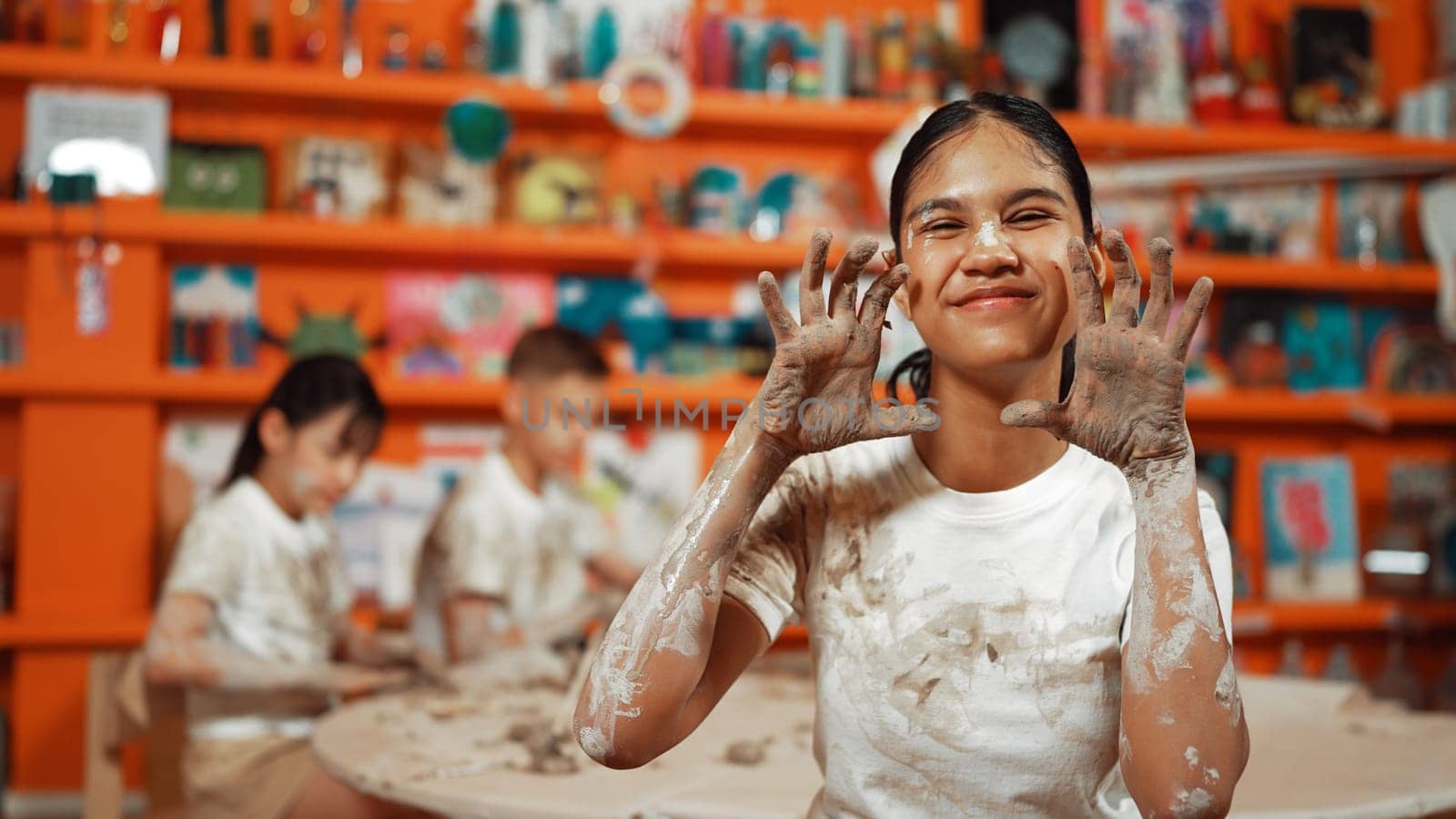 Happy caucasian girl pose at camera while diverse children modeling clay behind. Cute student wearing dirty shirt while looking at camera at workshop in art lesson. Blurring background. Edification.