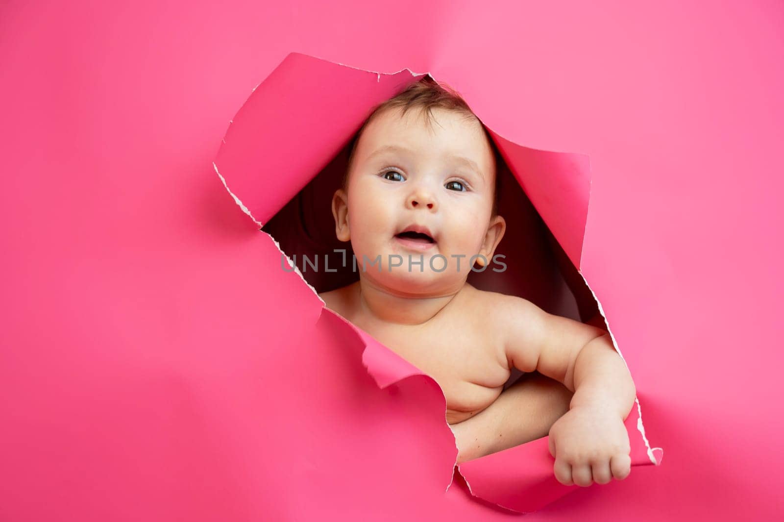 Cute Caucasian newborn baby boy peeks out of a hole in a paper pink background