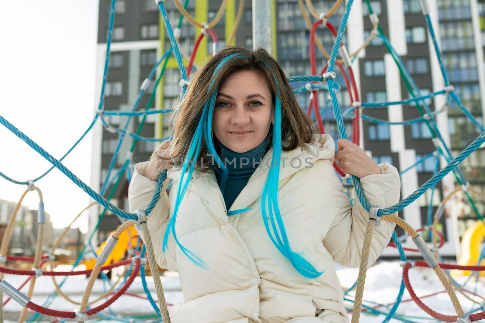 A woman with vibrant blue hair is sitting on a colorful playground structure. She looks relaxed as she enjoys the fresh air and the playful surroundings of the playground.