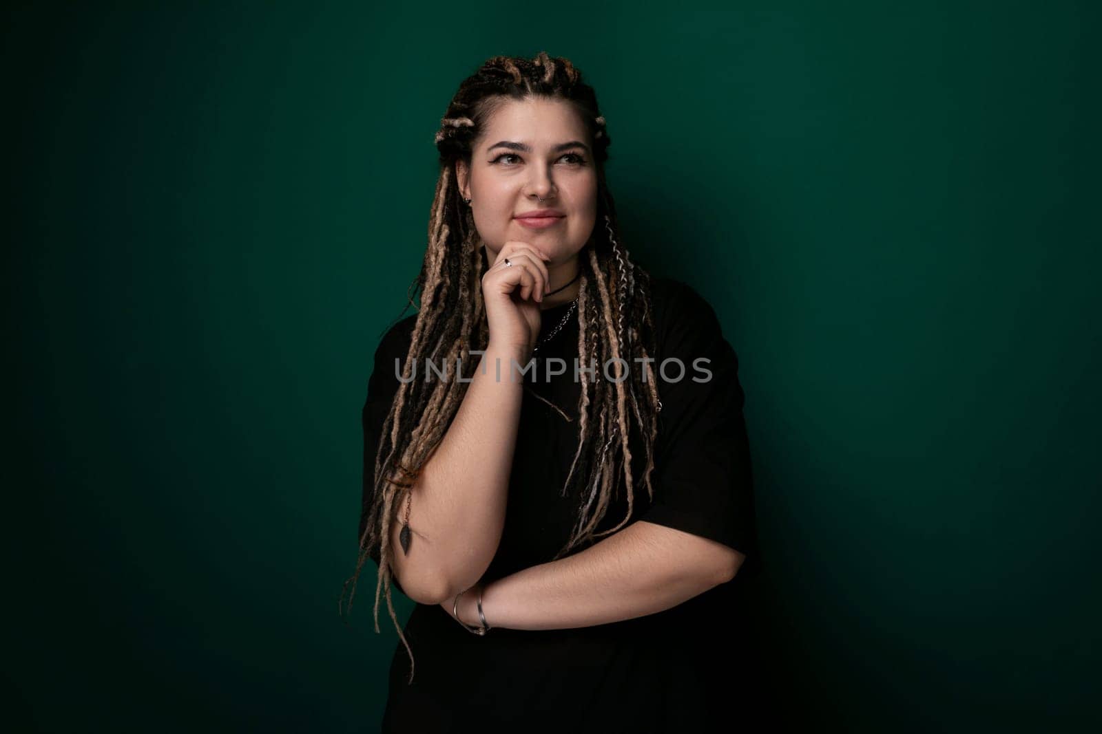 A woman with dreadlocks stands confidently in front of a vibrant green wall. Her unique hairstyle contrasts with the bold color behind her, creating a striking visual composition.