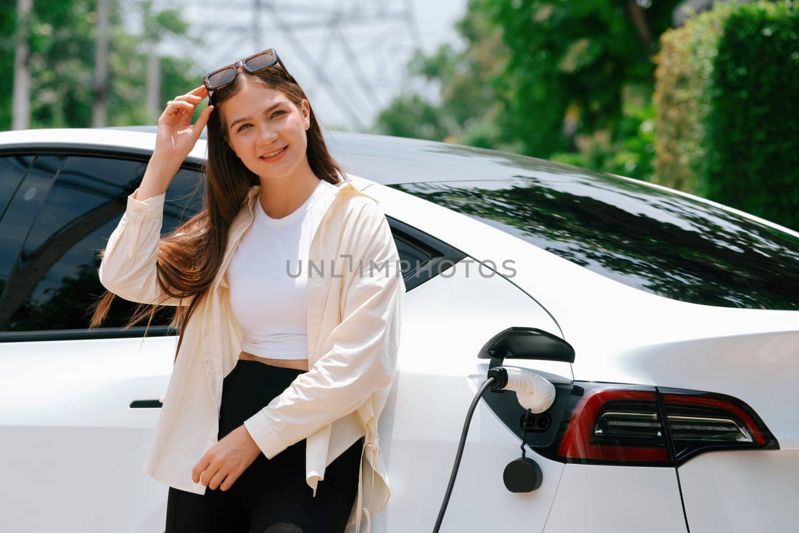 Young woman recharge her EV electric vehicle at green city park parking lot. Urban sustainability lifestyle for environmental friendly EV car with battery charging station. Expedient