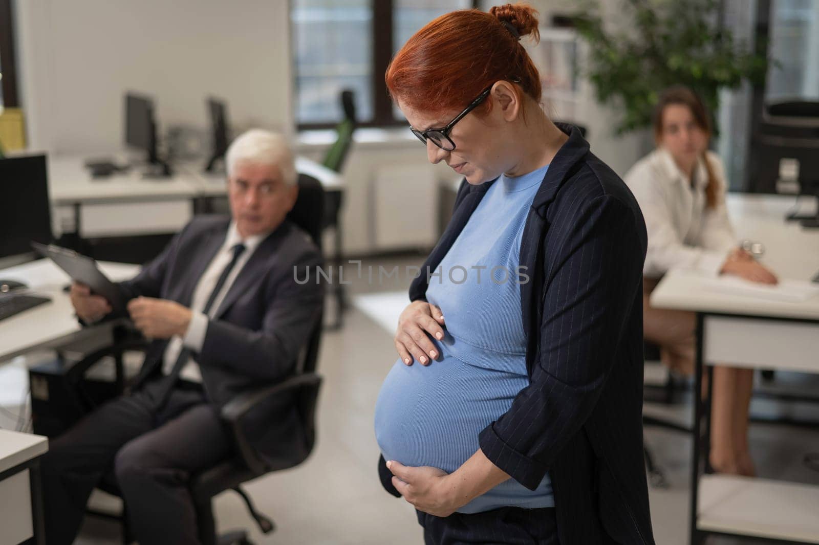 A pregnant woman suffers from pain and holds her stomach while standing in the middle of the office next to her colleagues. by mrwed54