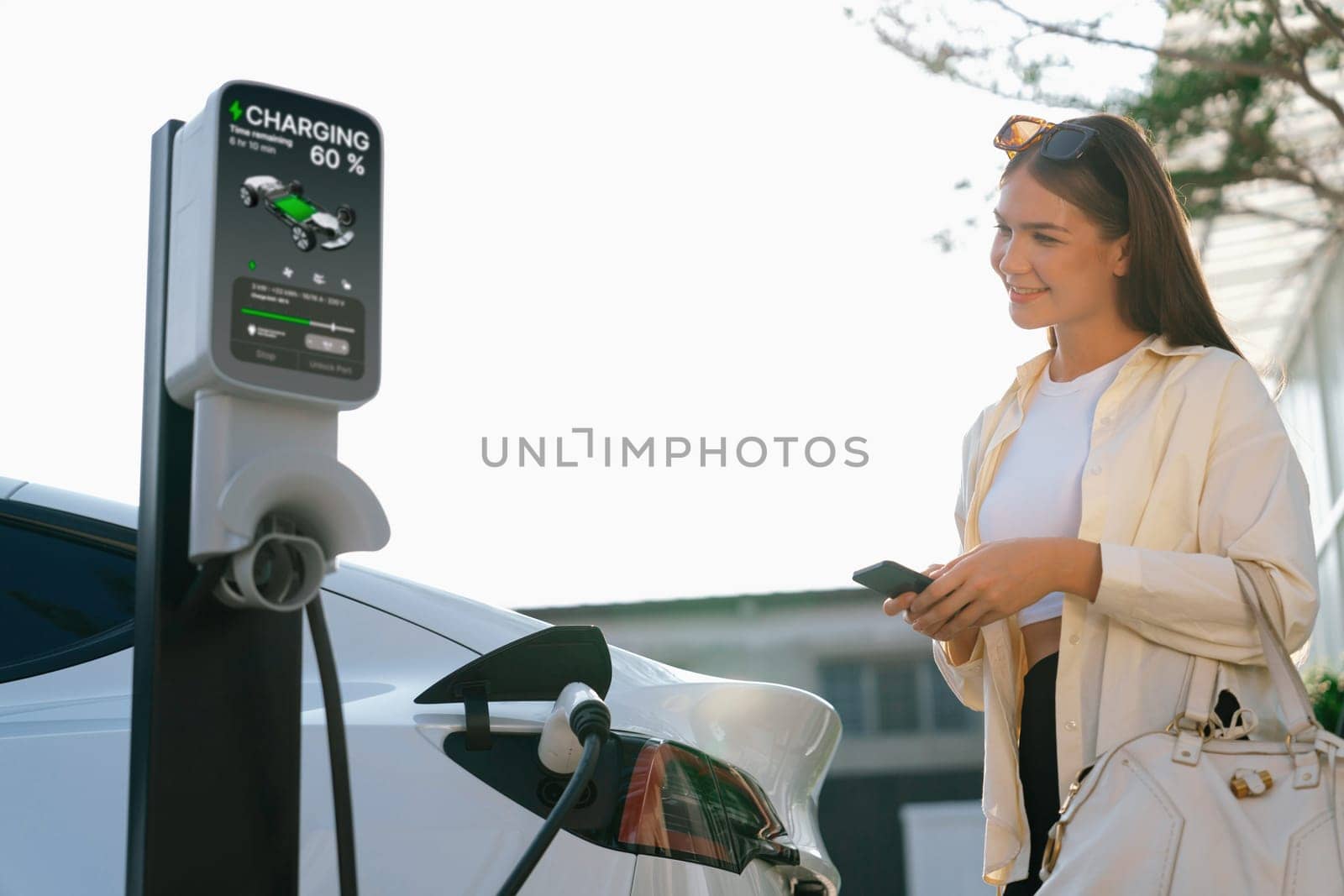 Young woman holding shopping bag and use smartphone to pay for electricity for recharging EV car battery from charging station at city mall parking lot. Modern woman go shopping by eco car. Expedient