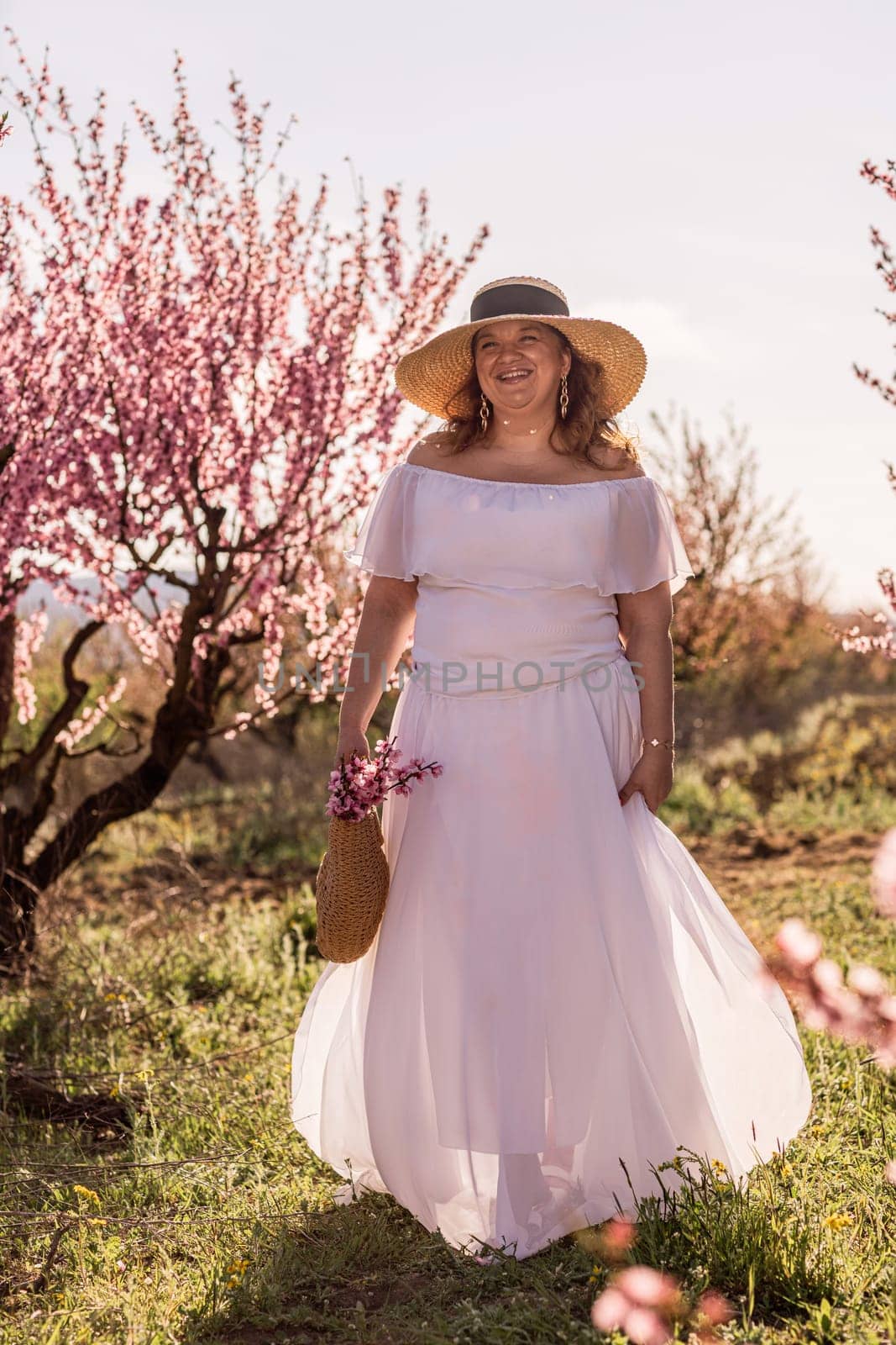 Woman blooming peach orchard. Against the backdrop of a picturesque peach orchard, a woman in a long white dress and hat enjoys a peaceful walk in the park, surrounded by the beauty of nature