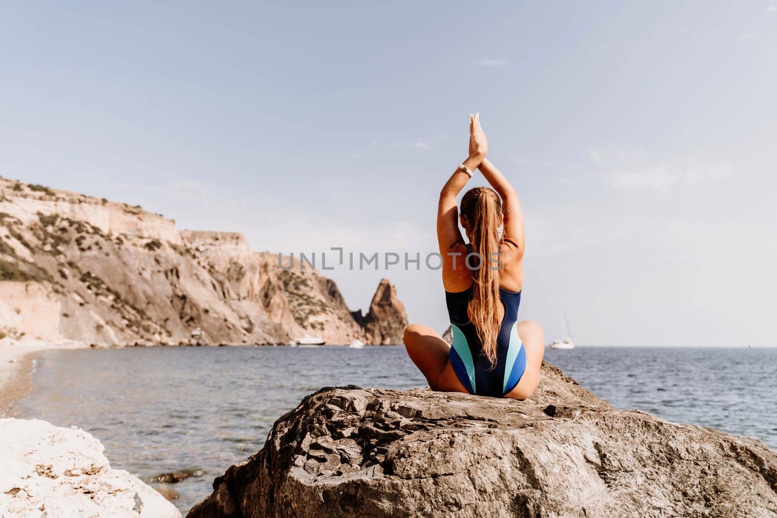Yoga on the beach. A happy woman meditating in a yoga pose on the beach, surrounded by the ocean and rock mountains, promoting a healthy lifestyle outdoors in nature, and inspiring fitness concept
