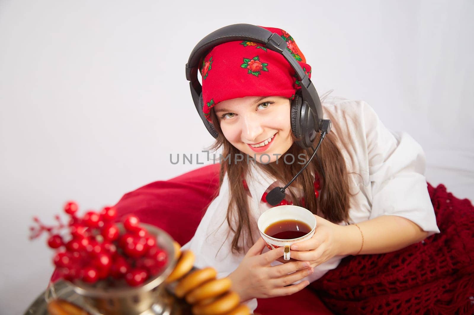 Young girl in red scarf and large headphones with microphone with samovar, bagels, cap of tea on Easter or Maslenitsa. Woman radio or television presenter, female telecom operator, blogger, Freelancer