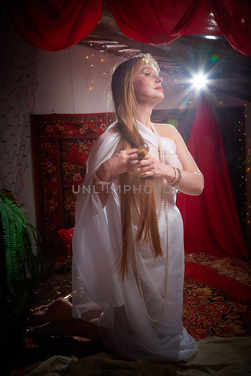 Beautiful European girl looking like Arab woman in red room with rich fabrics and carpets in sultan harem. Photo shoot of an oriental style odalisque. A model poses in sari as indian woman in india