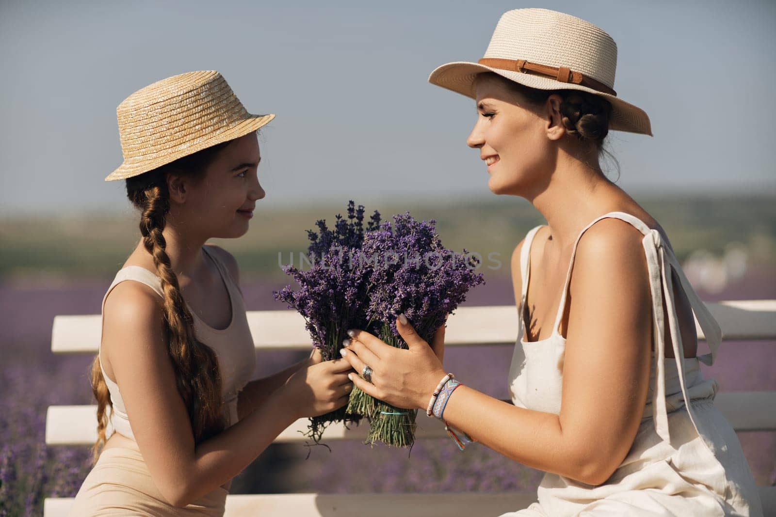 A woman and a child are sitting on a bench in a field of purple flowers lavande. The woman is holding a bouquet of flowers and the child is holding a bouquet of flowers as well by Matiunina