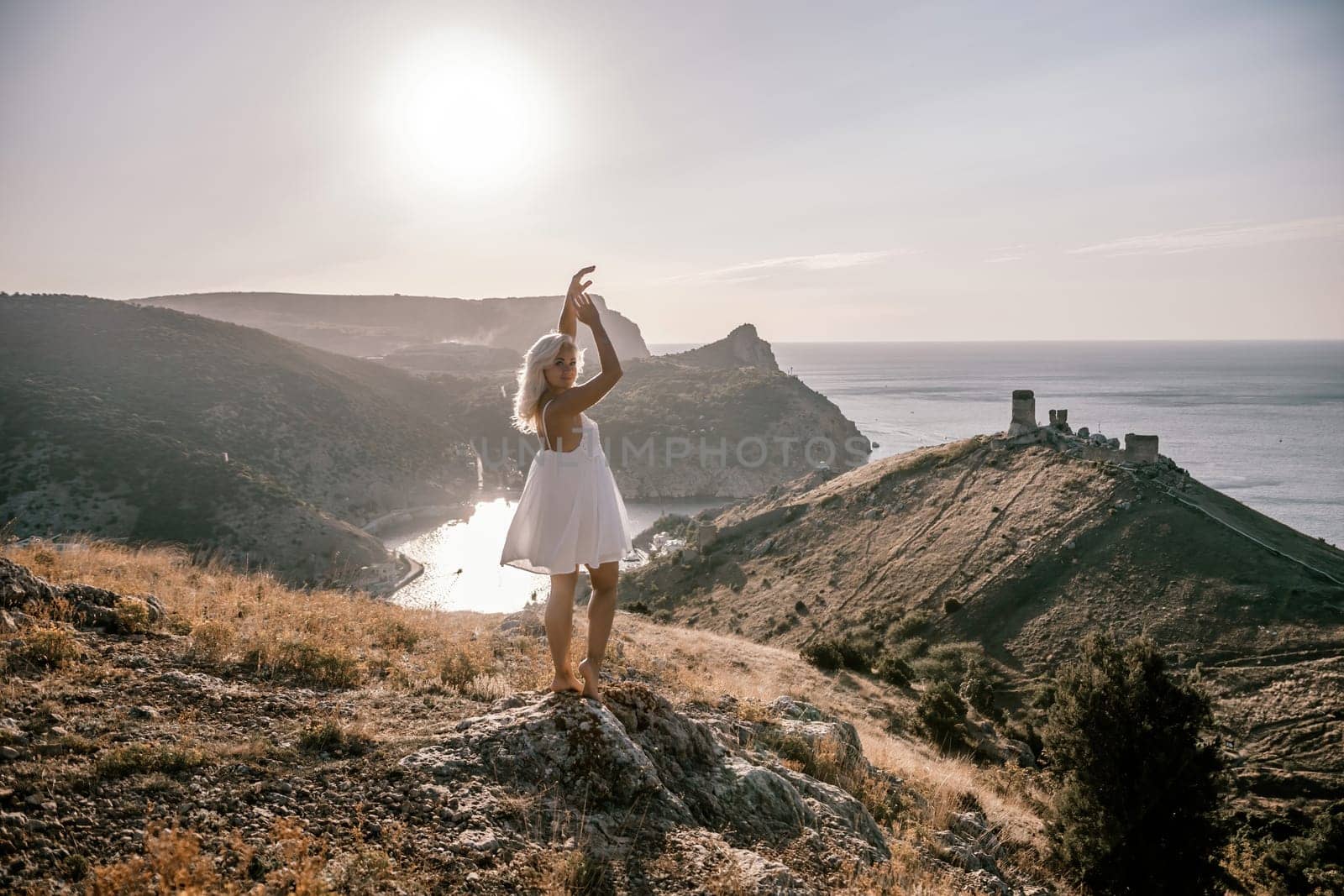 A woman stands on a hill overlooking a body of water. She is wearing a white dress and she is enjoying the view. by Matiunina