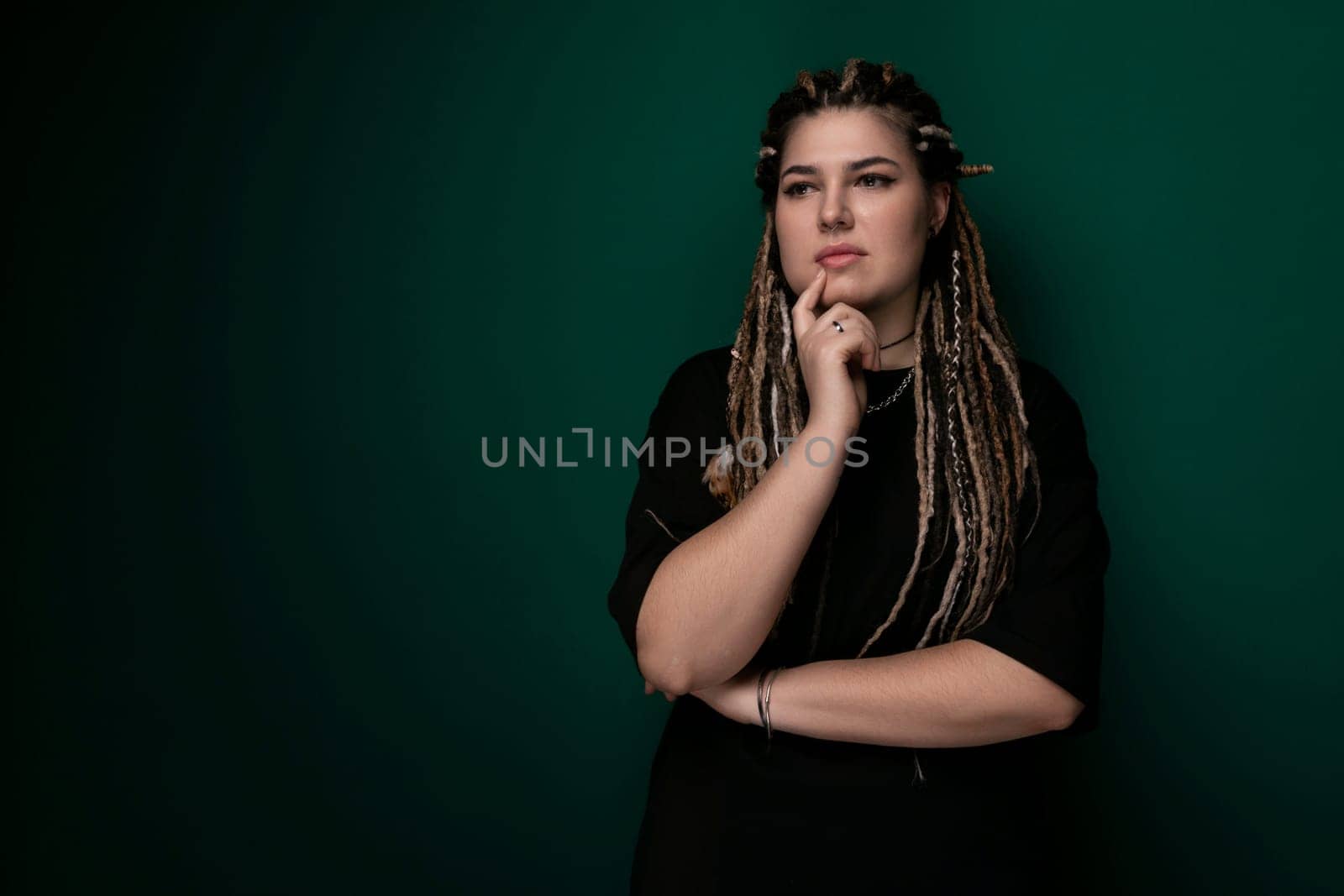 A woman with dreadlocks is standing in front of a green wall. She is looking directly at the camera, appearing confident and composed.