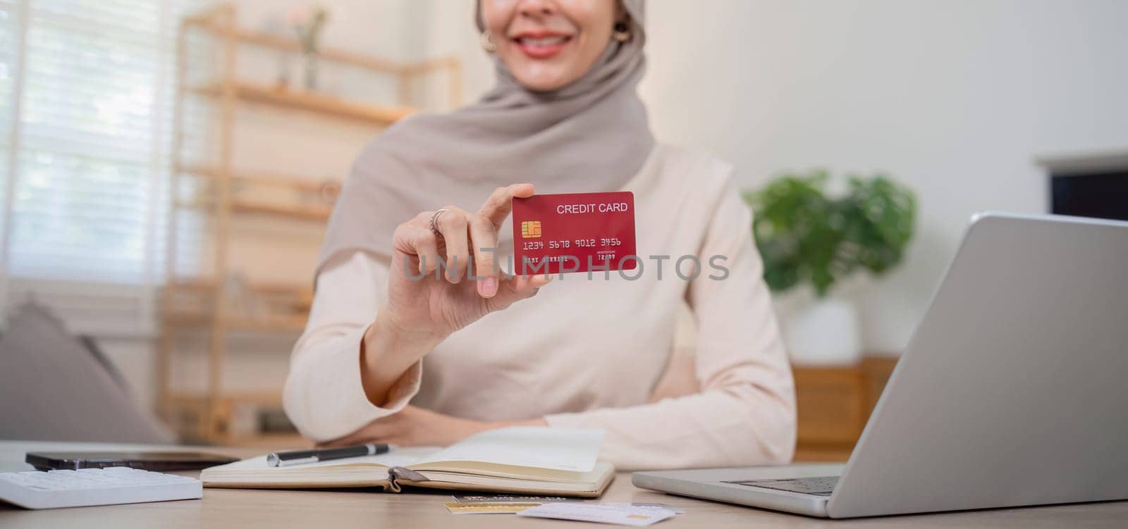 A young Muslim woman wearing a hijab sat contentedly shopping online and holding a credit card. by wichayada