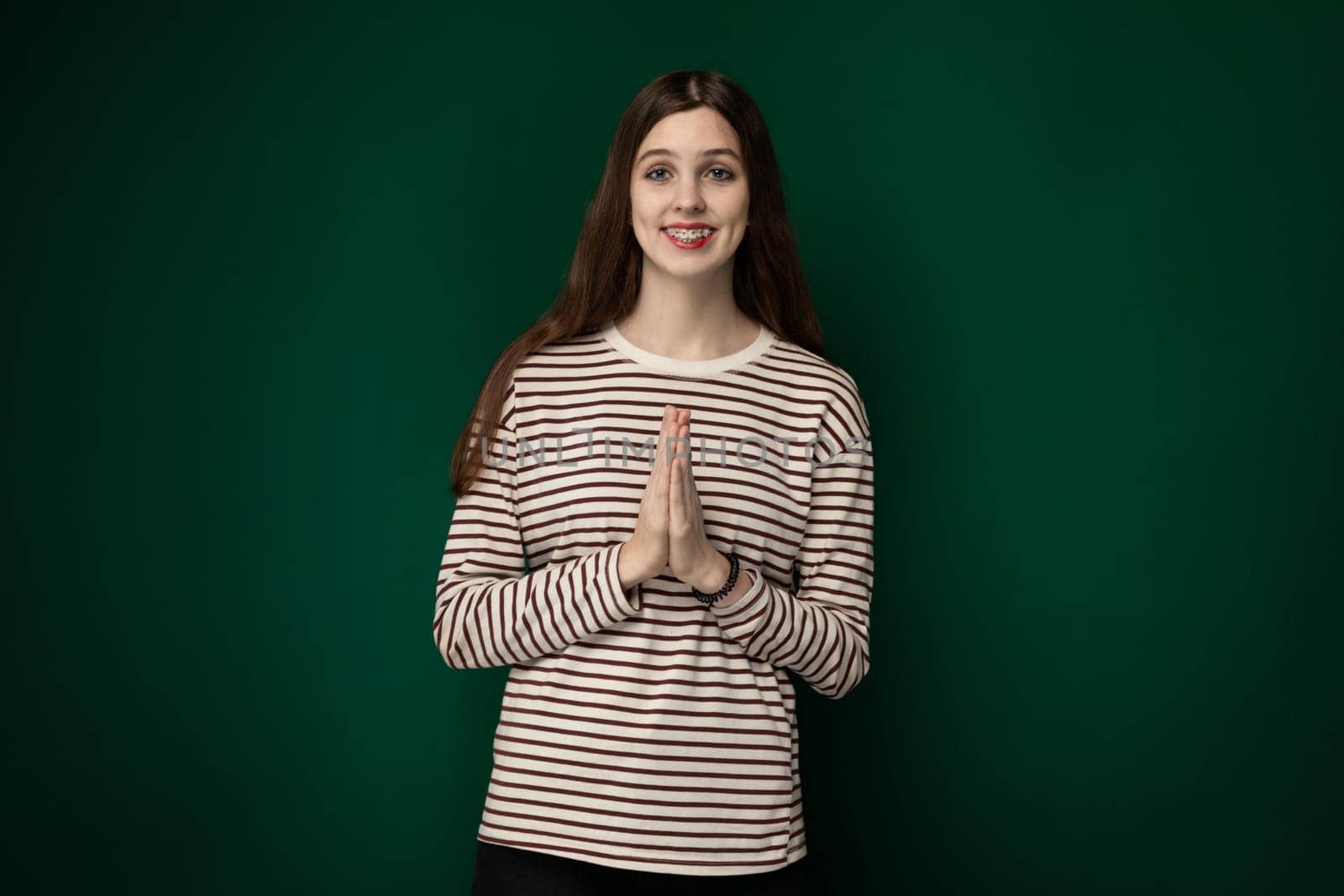 A woman is standing upright in front of a vibrant green wall, gazing directly at the camera. She is wearing casual attire and appears confident in posture. The simplicity of the scene highlights the contrast between the neutral backdrop and the vivid green hue.