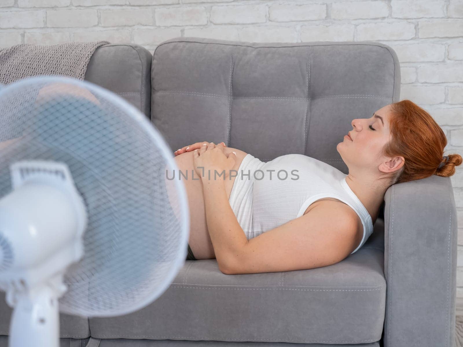 Pregnant woman suffers from heat and lies on the sofa near the electric fan