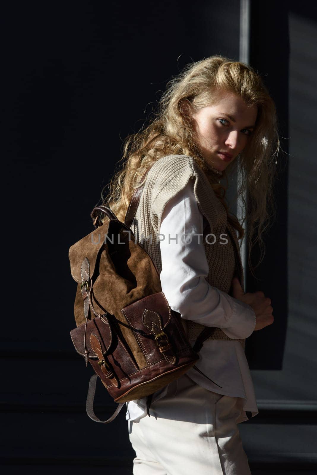 A woman holds brown leather backpack. Model wearing stylish knitted vest, white shirt and classic trousers.