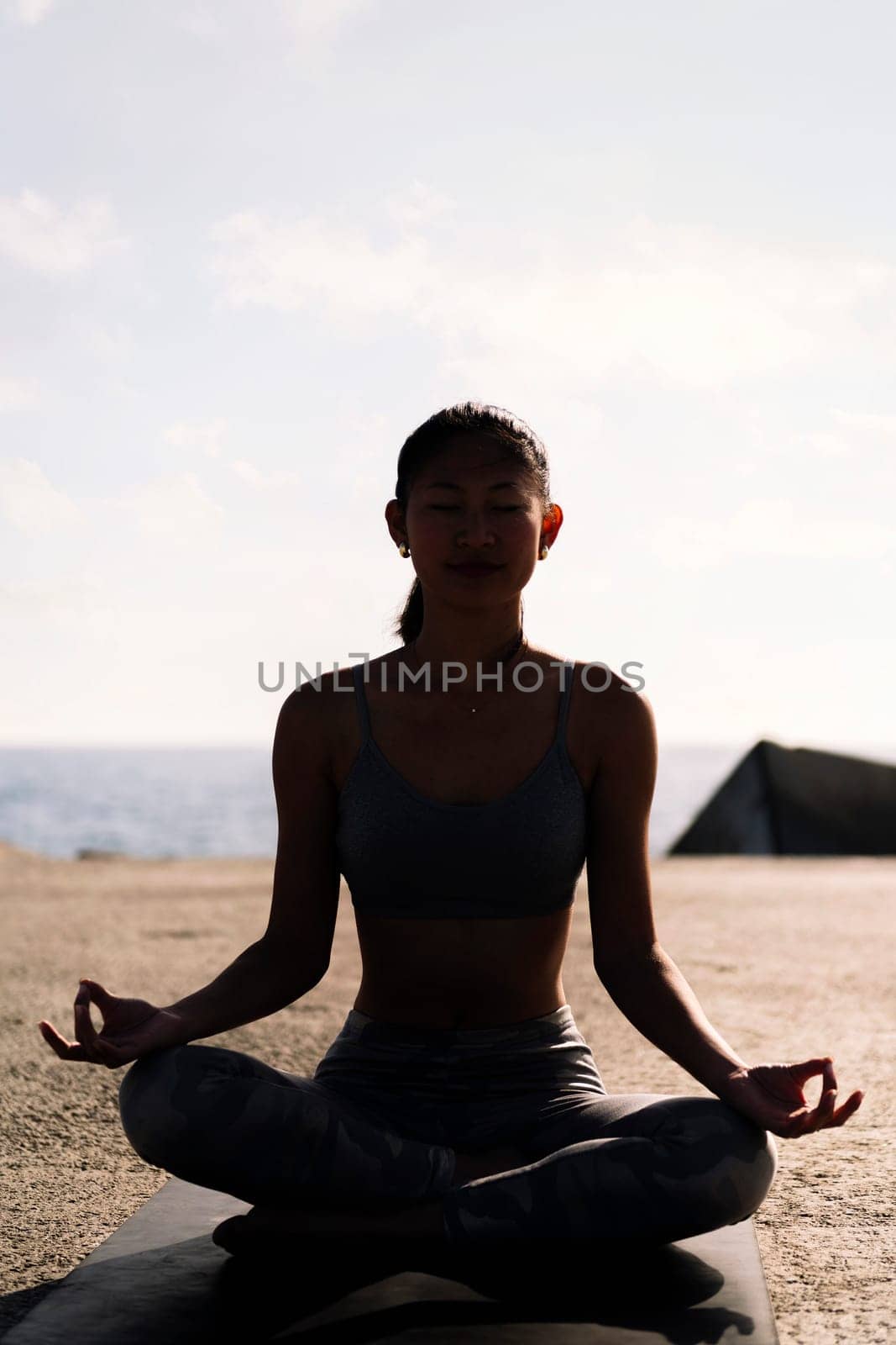 backlight of young woman doing meditation by the sea sitting with legs crossed, concept of mental relaxation and healthy lifestyle