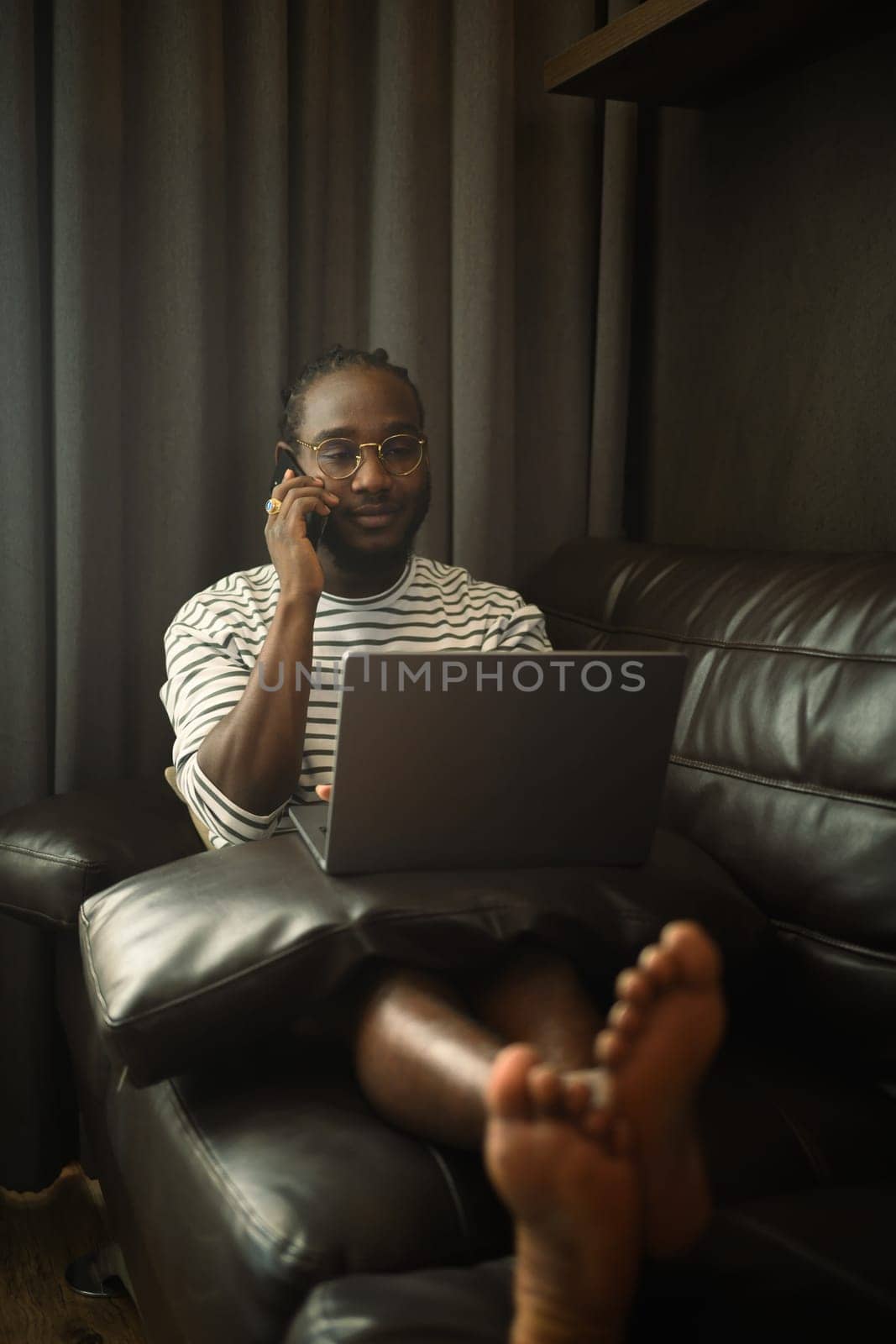 African American man talking mobile phone and using laptop on couch by prathanchorruangsak