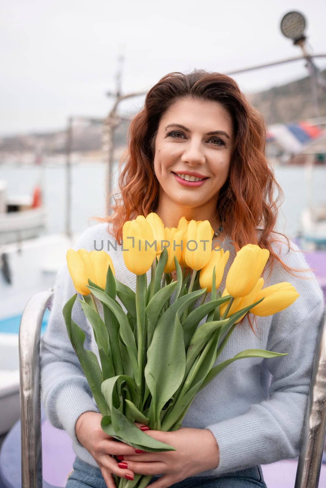 Woman holds yellow tulips in harbor with boats docked in the background., overcast day, yellow sweater, mountains by Matiunina