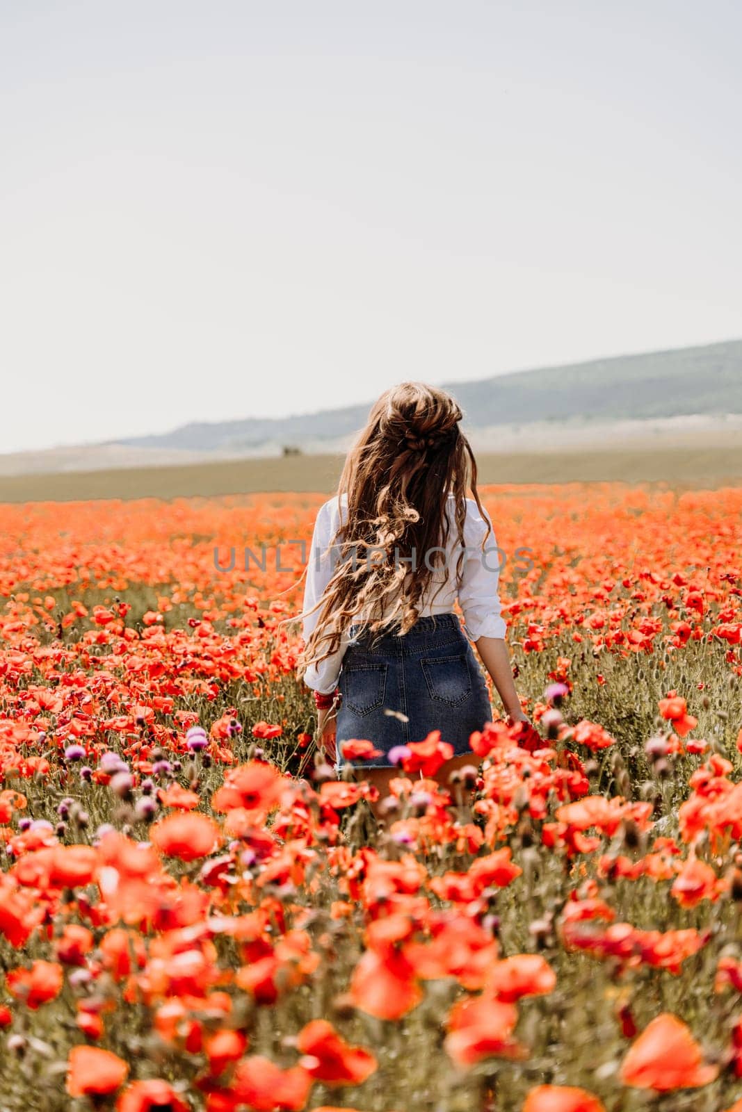 Happy woman in a poppy field in a white shirt and denim skirt with a wreath of poppies on her head posing and enjoying the poppy field. by Matiunina