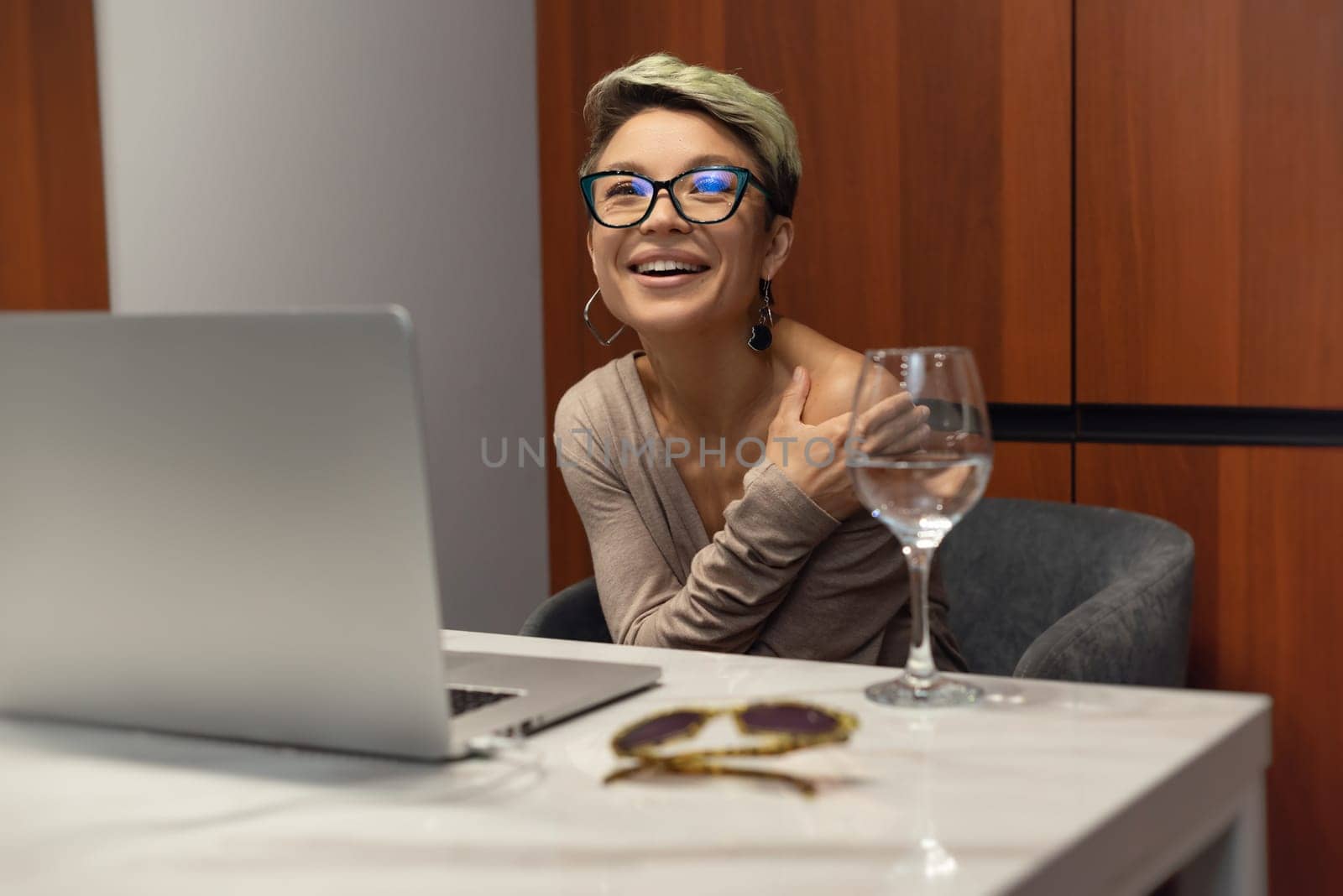 girl with short hair and glasses indoors at a laptop laughs and smiles emotionally and cheerfully while working and chatting