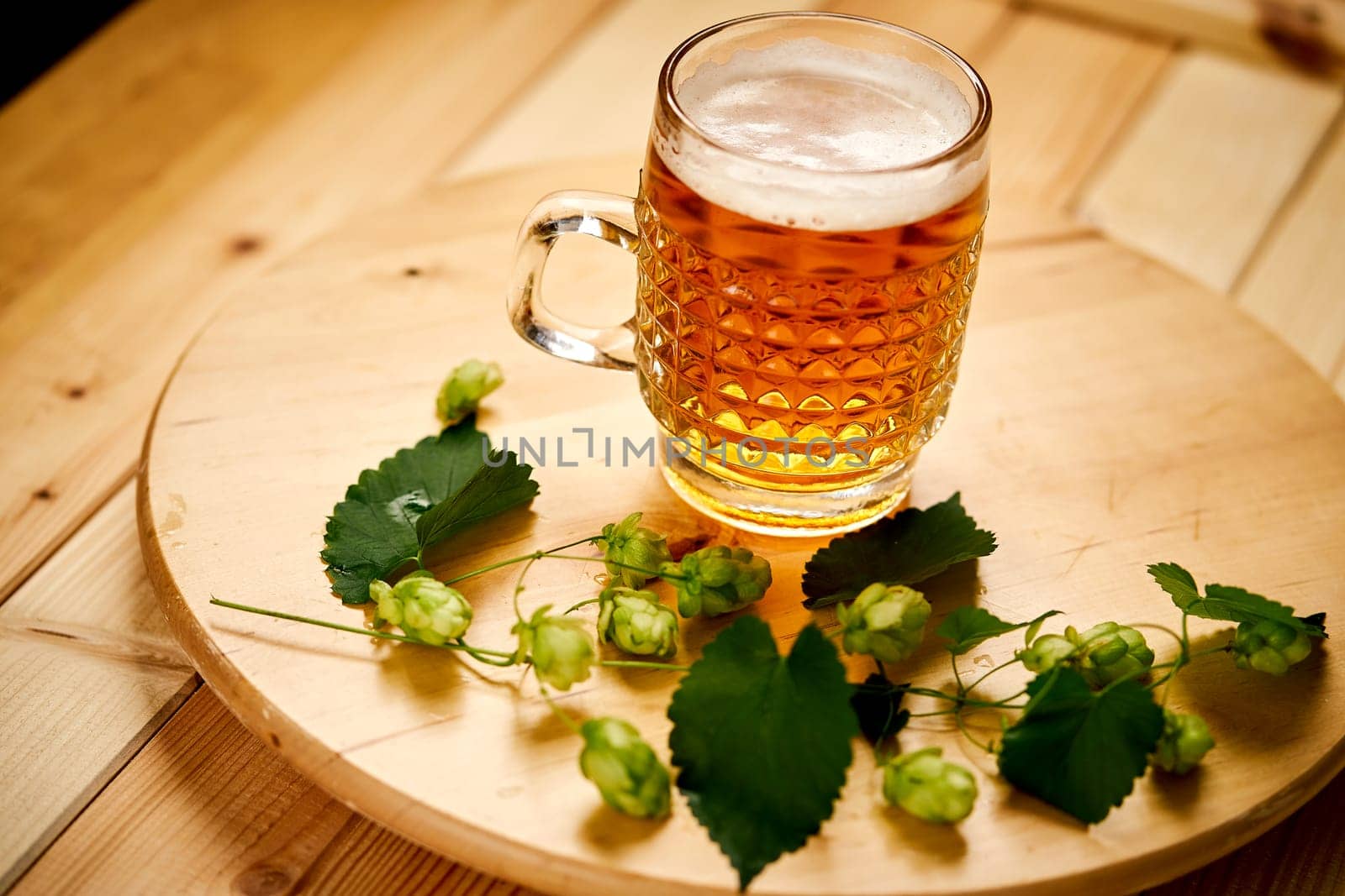 Large pint of beer with foam and sprigs of hops on bright wooden table