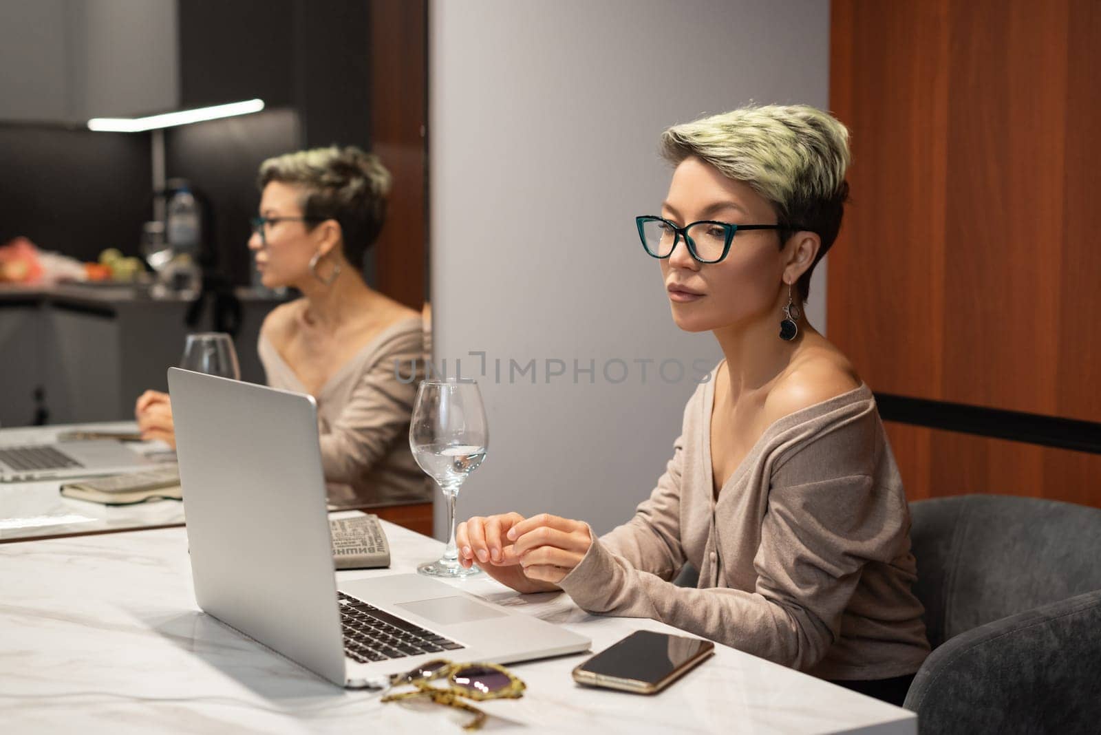 a beautiful girl with short hair and glasses is sitting indoors at a laptop chatting and working online by Rotozey
