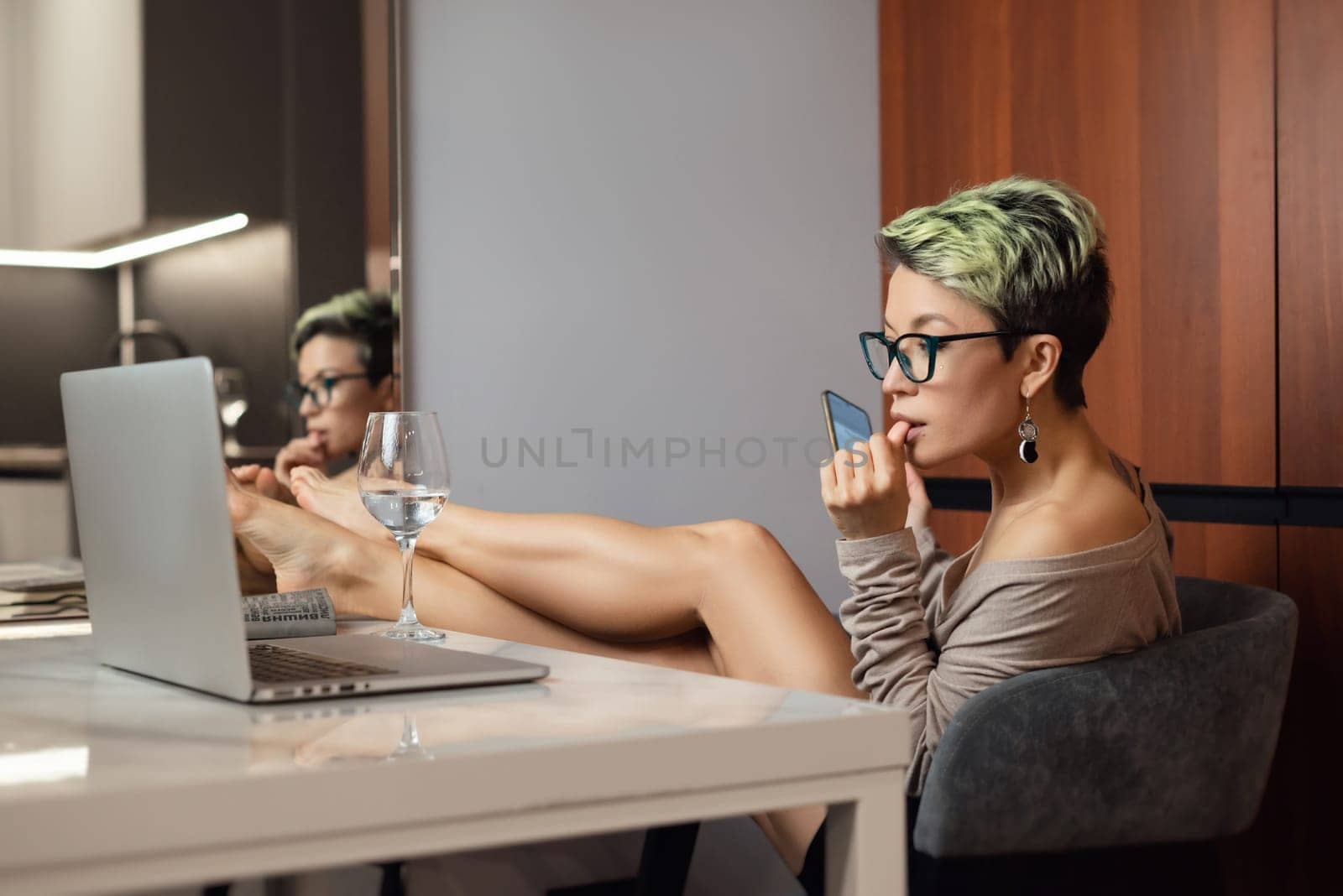 a beautiful girl with short hair and glasses is sitting indoors at a laptop, with her feet on the table, chatting and working online by Rotozey