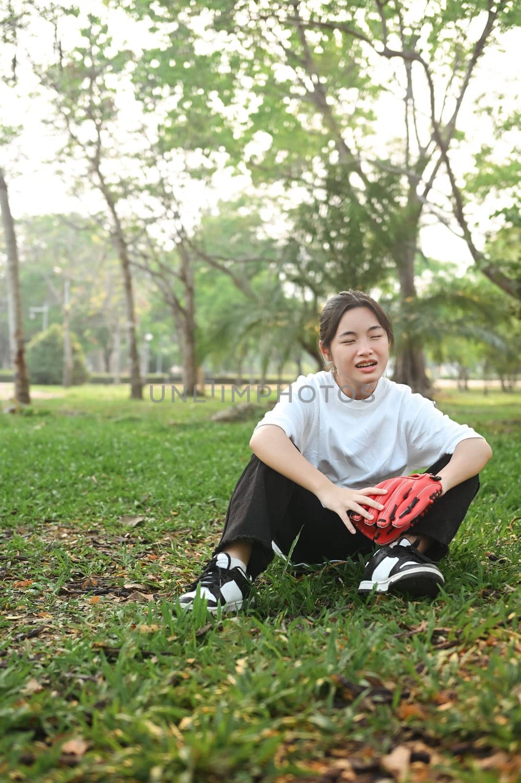 Happy teenage girl wearing leather baseball glove sitting on green lawn during summer day by prathanchorruangsak