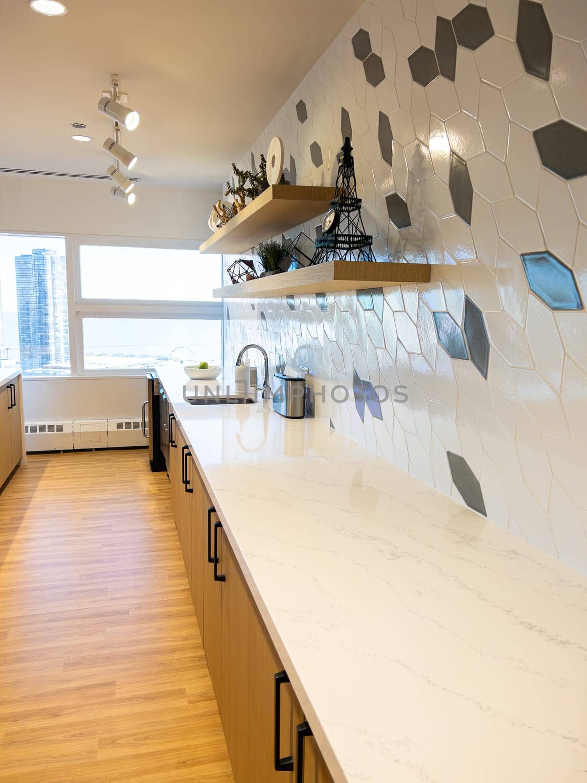 A compact bar area within a hotel's fitness center, featuring a striking geometric backsplash and floating wooden shelves, with a panoramic window offering views of the urban skyline.