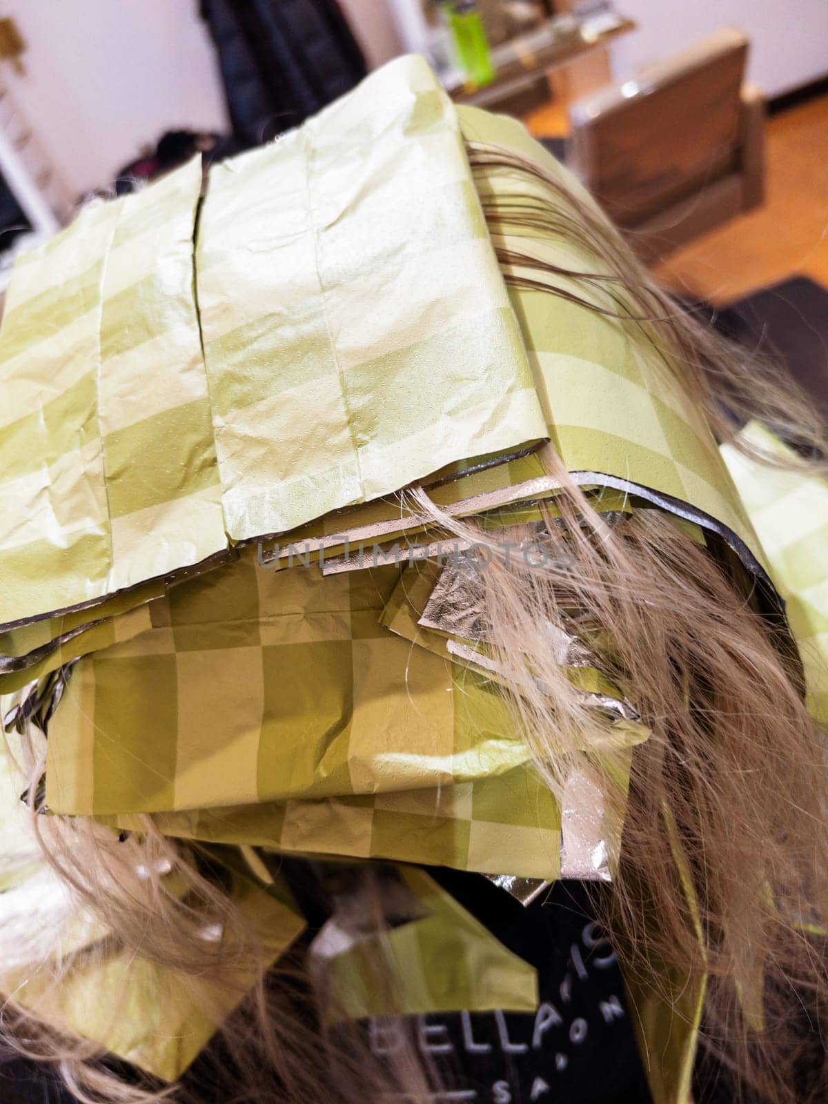 Strands of hair peek through a series of yellow foils meticulously arranged for a highlighting treatment. The salon's ambiance is reflected in the background, where focused stylists perform their craft, transforming hair with skill and precision.