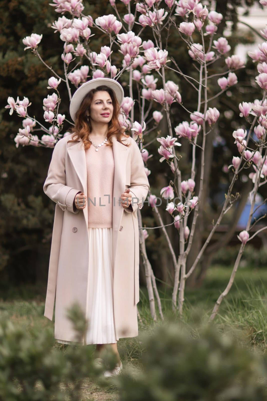 Woman magnolia flowers, surrounded by blossoming trees, hair down, white hat, wearing a light coat. Captured during spring, showcasing natural beauty and seasonal change