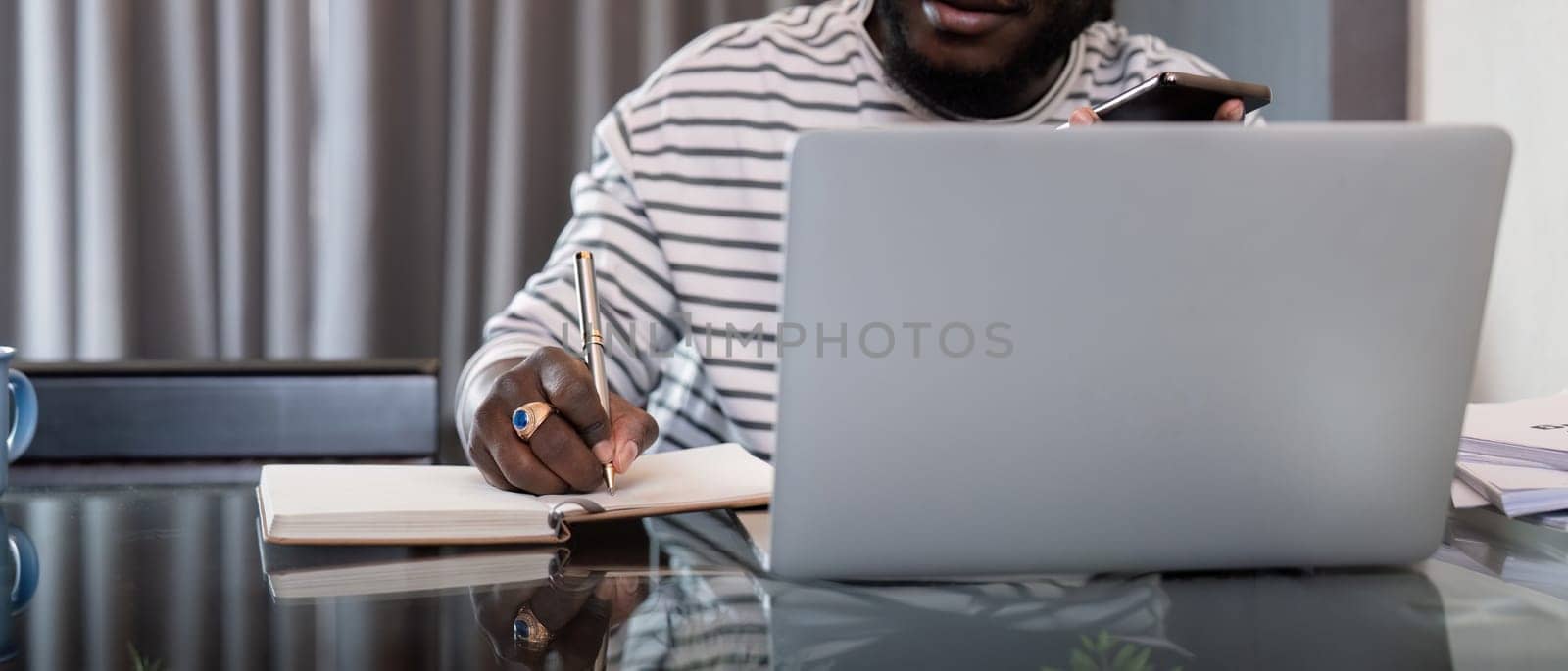 African American man working with laptop computer remote while sitting at glass table in living room. Black guy do freelance while taking notes work at home office by nateemee
