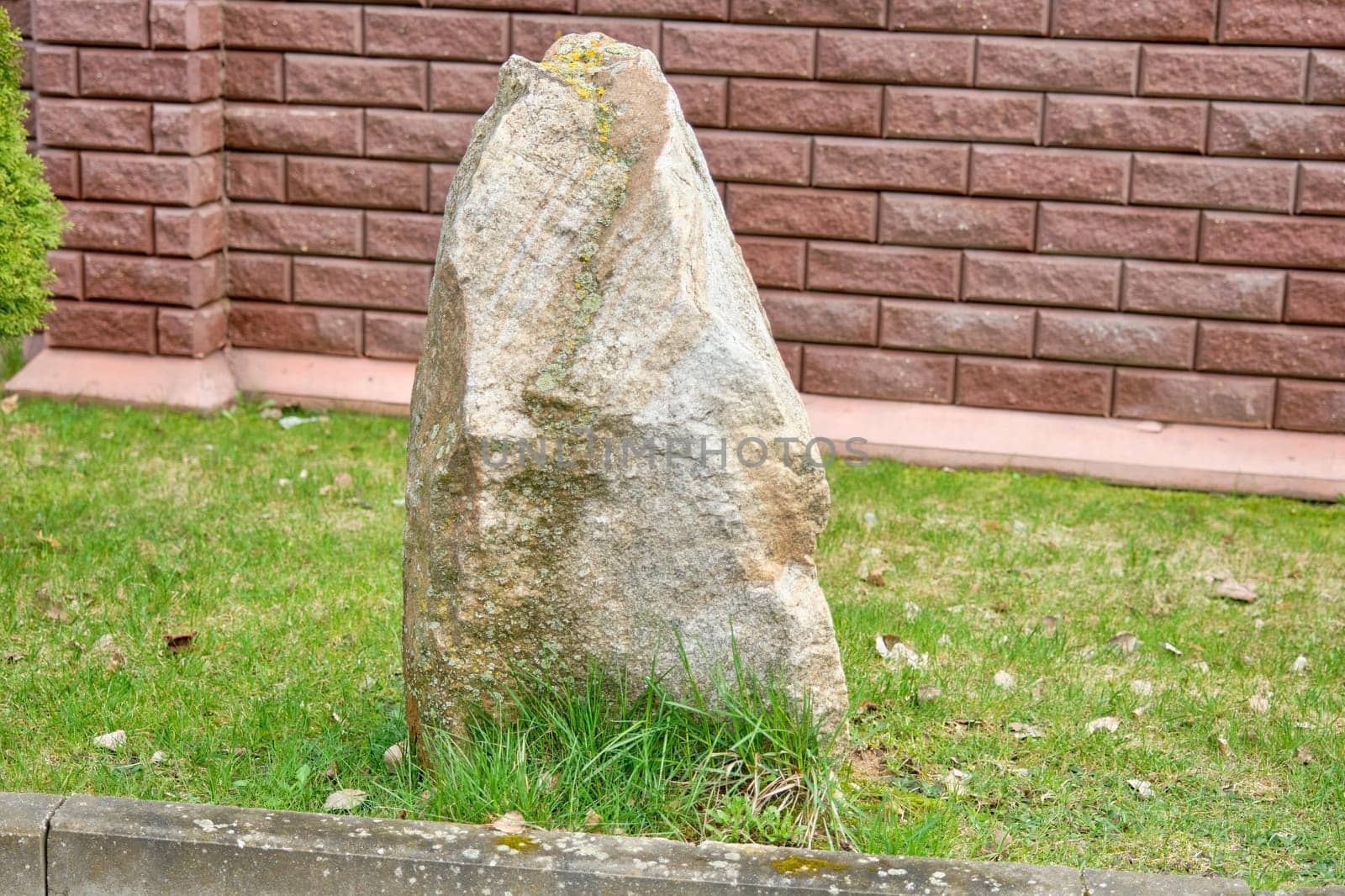 A large stone lies on the grass in front of a red brick fence by Ihar