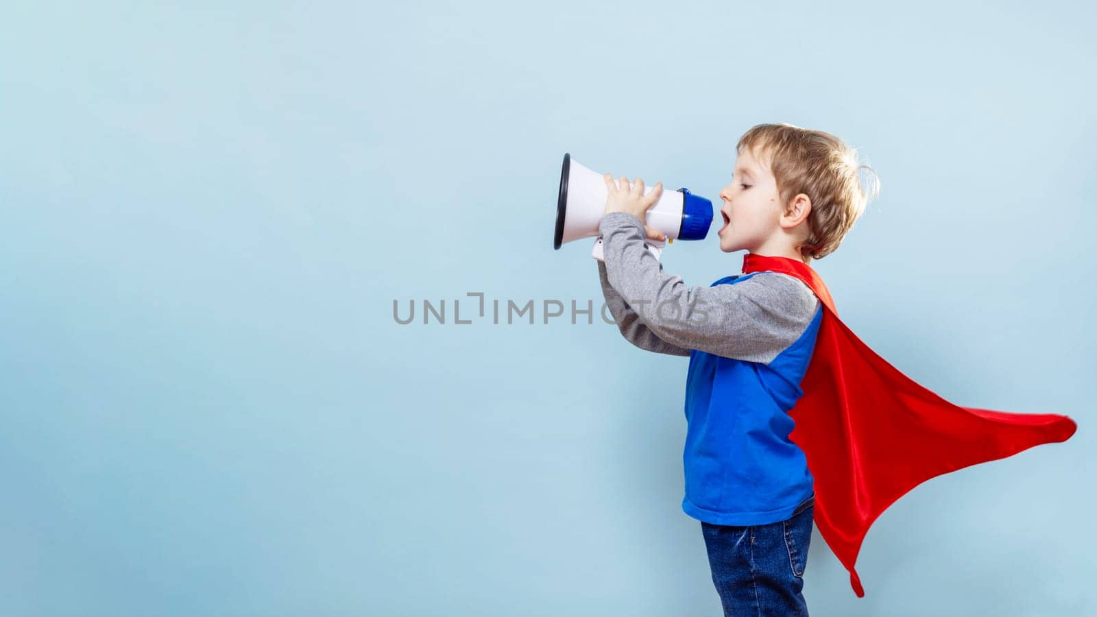 Young boy in superhero cape shouting through megaphone on blue background. Childhood play and imagination concept for poster design.
