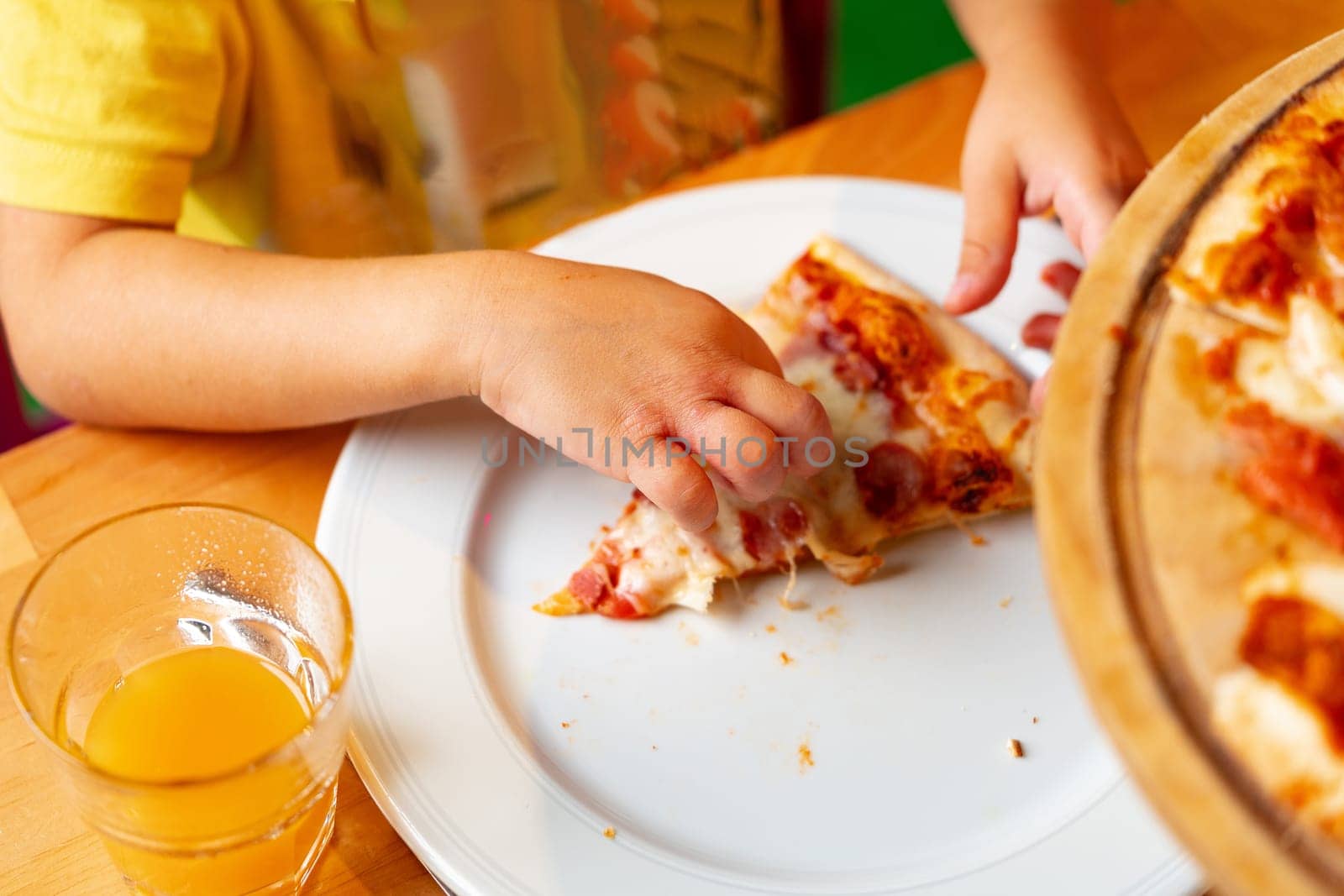 A child sitting at a table, holding a slice of pizza in their hand and taking a bite. The plate is placed in front of them, with pizza toppings visible. The child appears to be enjoying the food.