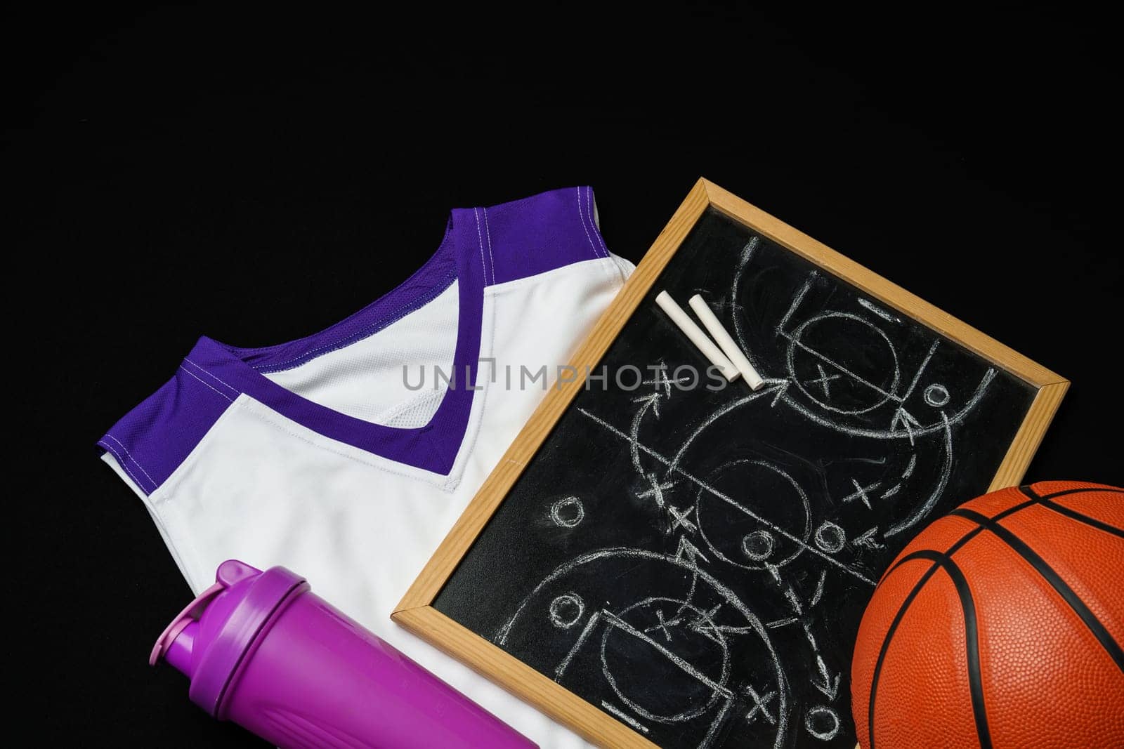 A basketball, along with a strategic play diagrammed on a chalkboard, rests near a folded jersey and a sports bottle. The equipment is arranged on a dark surface, suggesting preparation for a game or a coaching session in progress.