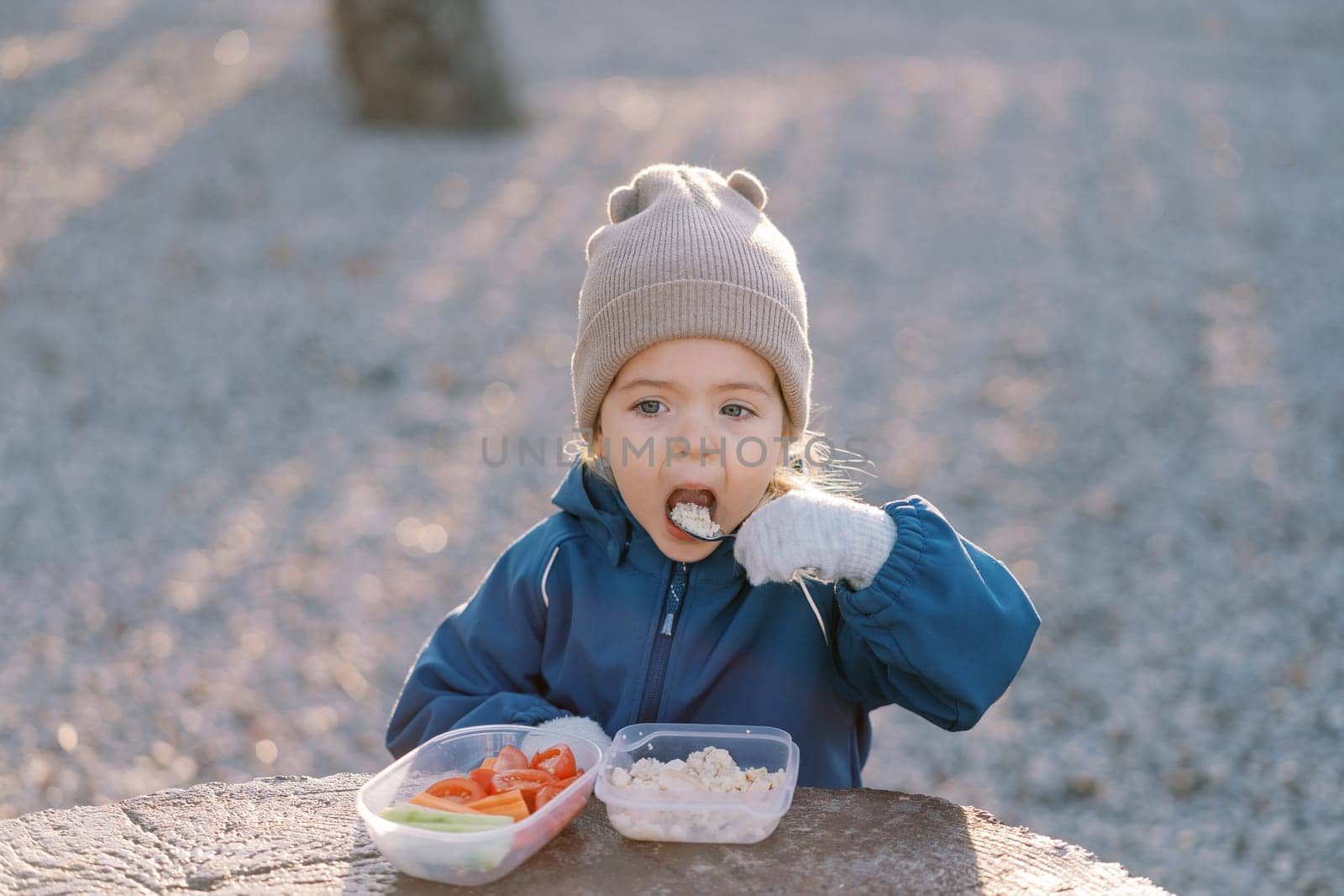 Little girl eats porridge with a spoon while standing behind a large tree stump with a lunchbox. High quality photo