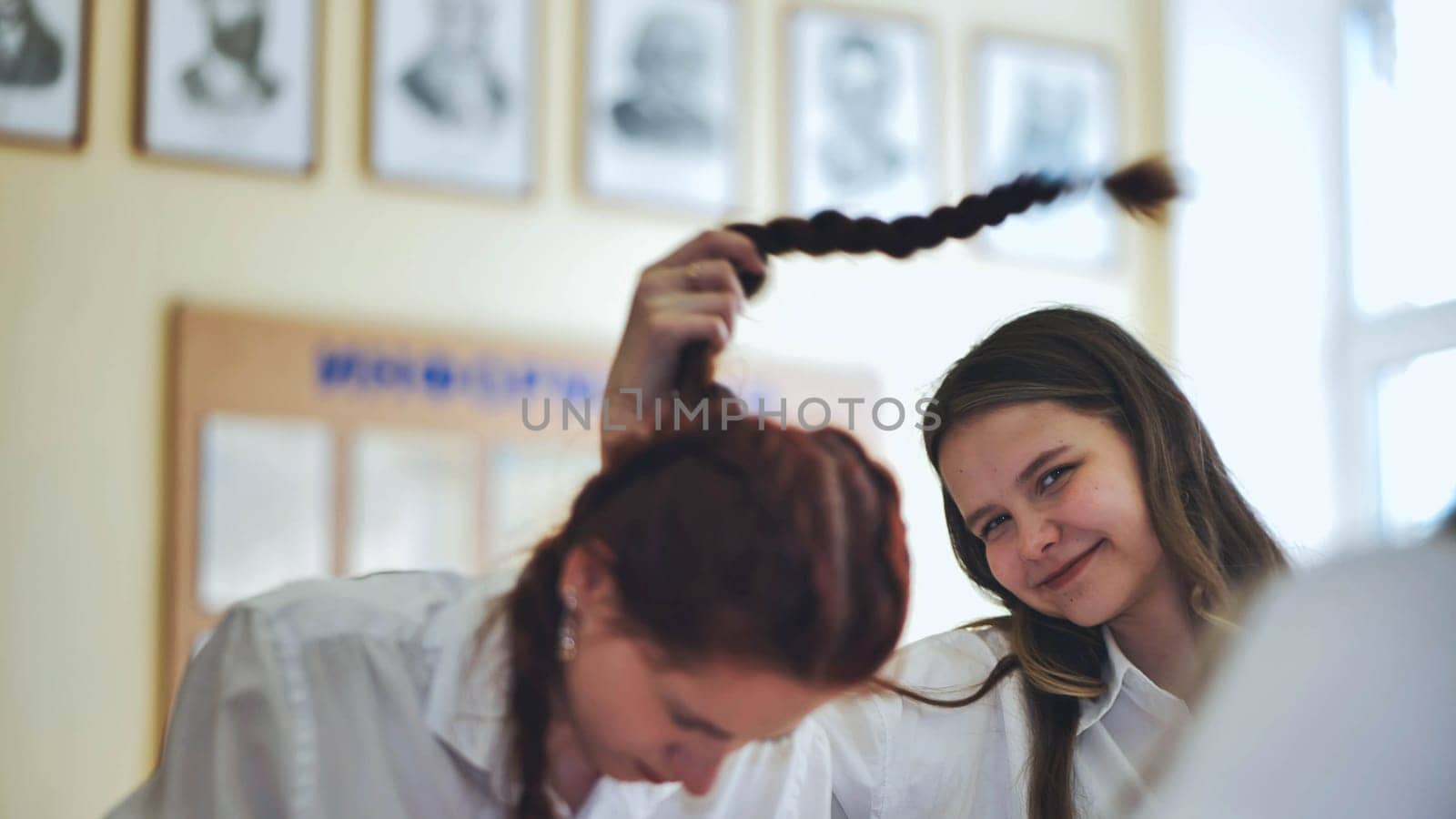 Fun schoolgirls in the classroom. Girl playing with her friend's pigtail. by DovidPro