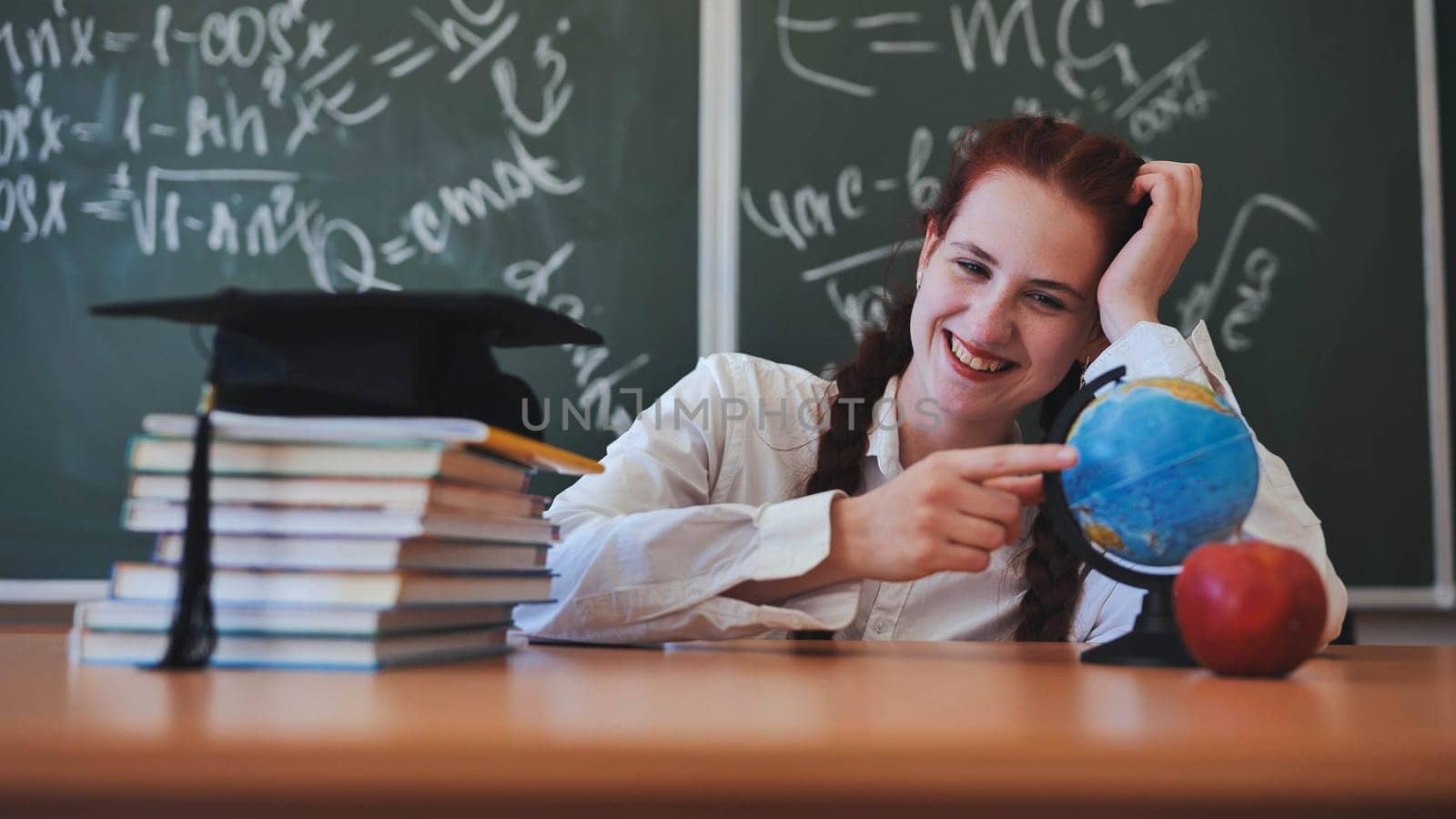 A red-haired high school senior poses against a backdrop of books, a globe and a graduation cap