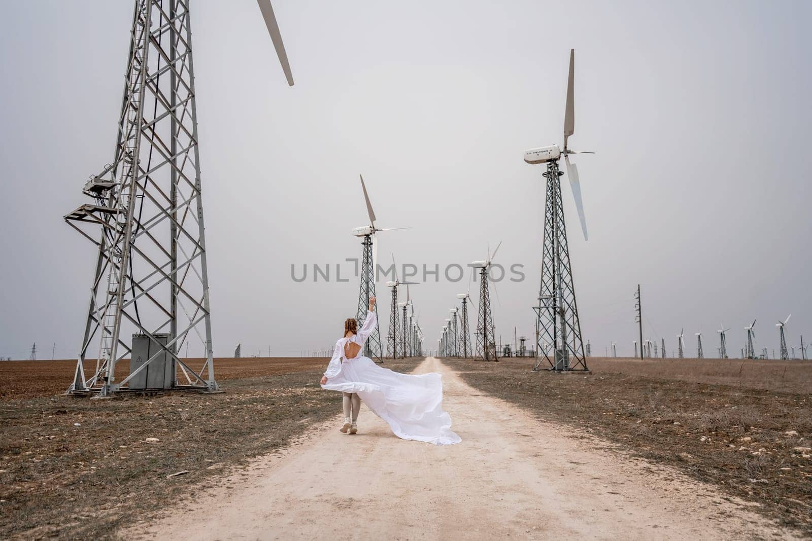 A woman in a white dress is walking down a dirt road in front of a row of wind turbines. by Matiunina