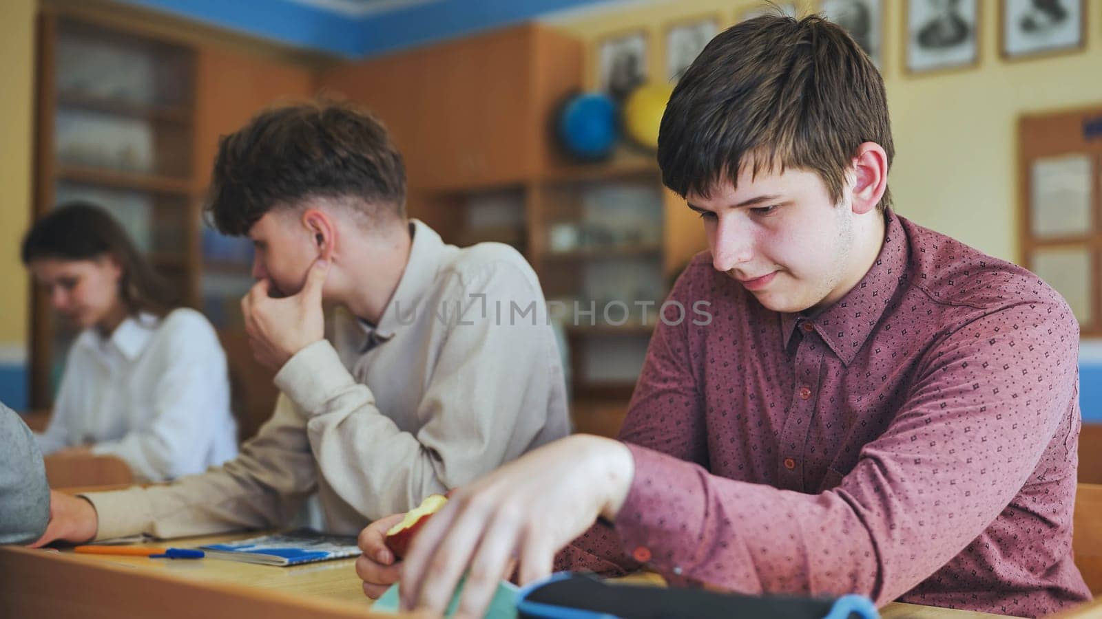 Schoolboys at a desk during class. The boy eats an apple. by DovidPro