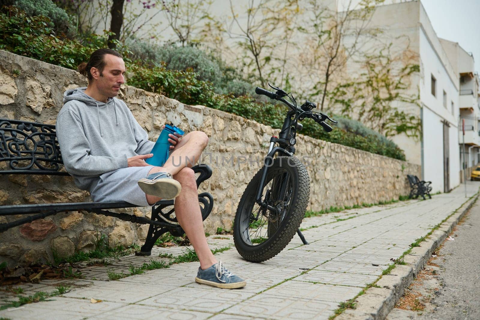 Young adult sportsman, athletic man cyclist relaxing when riding electric bike in the city, holding a bottle of water while sitting on the bench. Active people. Urban lifestyle. Eco-friendly transport by artgf