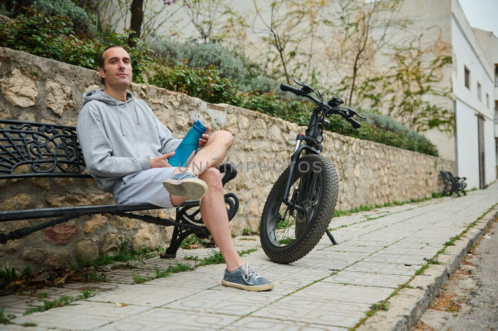 Young Caucasian man in gray hoodie and sports shorts, sitting on a bench near his rented electric bike, relaxing in the urban environment by artgf