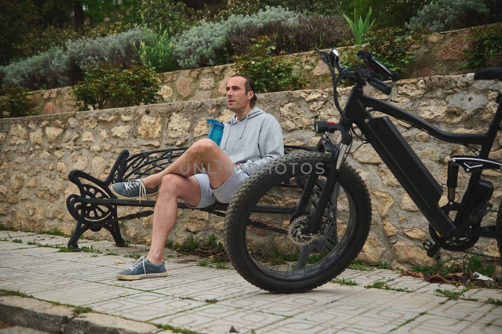 Handsome young cyclist man in sportswear, drinking water from sports bottle in the city, sitting on a bench, relaxing after riding an eco-friendly e-bike motorbike. Electric bicycle in the foreground