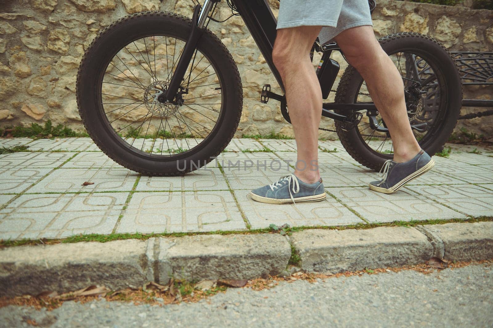 Cropped view legs of muscular sporty man pushing an electric mountain bike in the city. People. Healthy active lifestyle. Sport. Adventure travel on bicycle. Using el-bicycle as eco-friendly transport