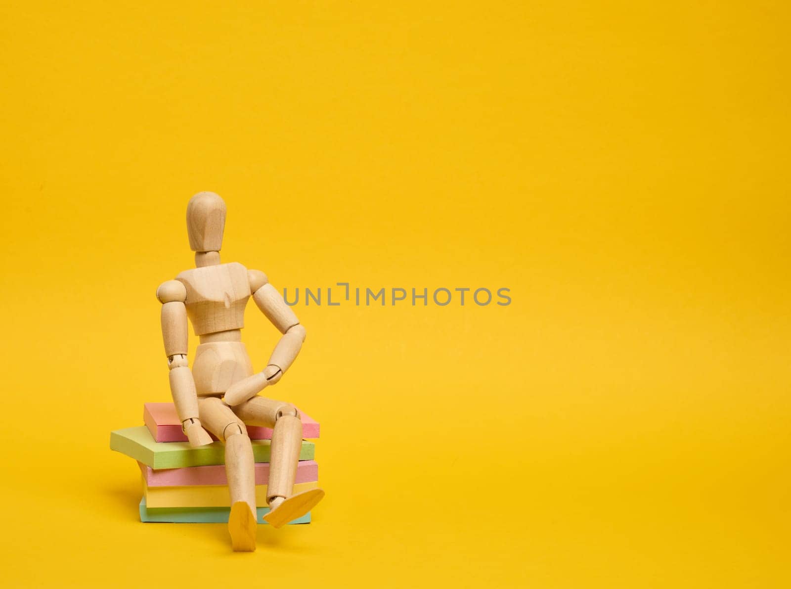 A wooden puppet doll sits on a stack of paper multi-colored stickers on a yellow background,  copy space